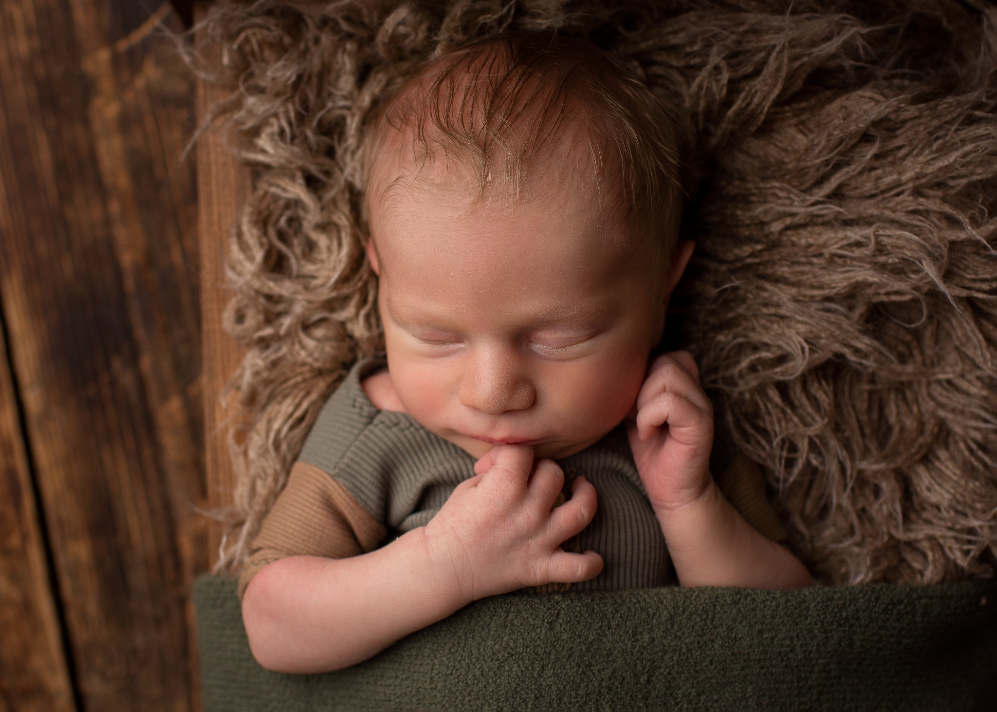Śpiący noworodek w zielonym ubranku, ułożony na brązowym futrze na tle drewnianych desek.