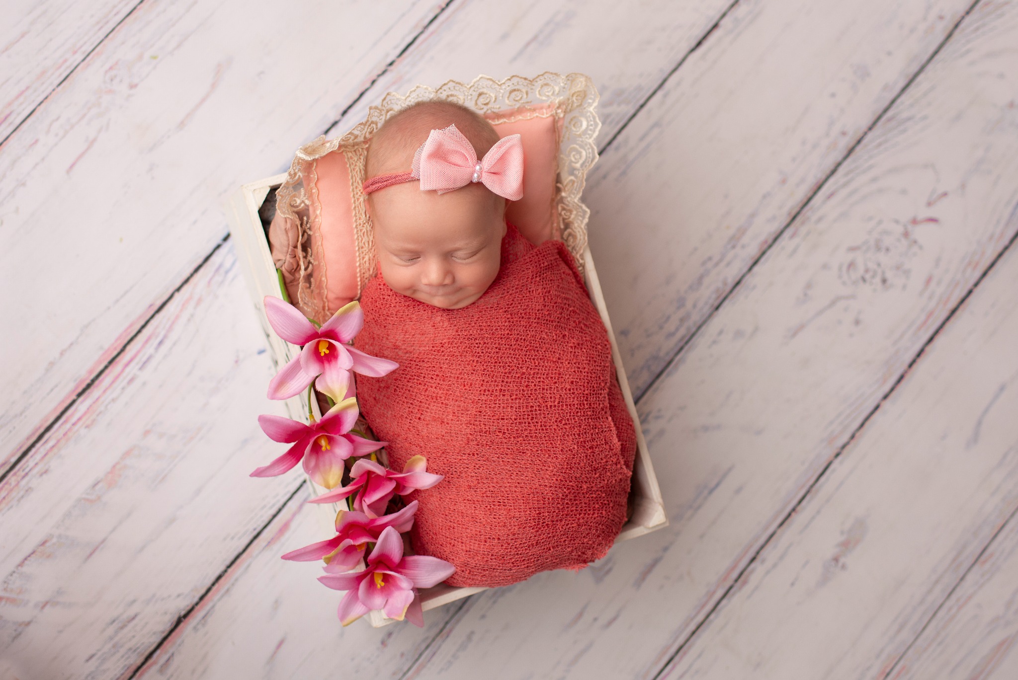 Noworodek zawinięty w koralowy kocyk, z różową kokardką na głowie, śpiący w białej drewnianej skrzynce z koronkowym obramowaniem i dekoracją z różowych orchidei na tle białych desek.
