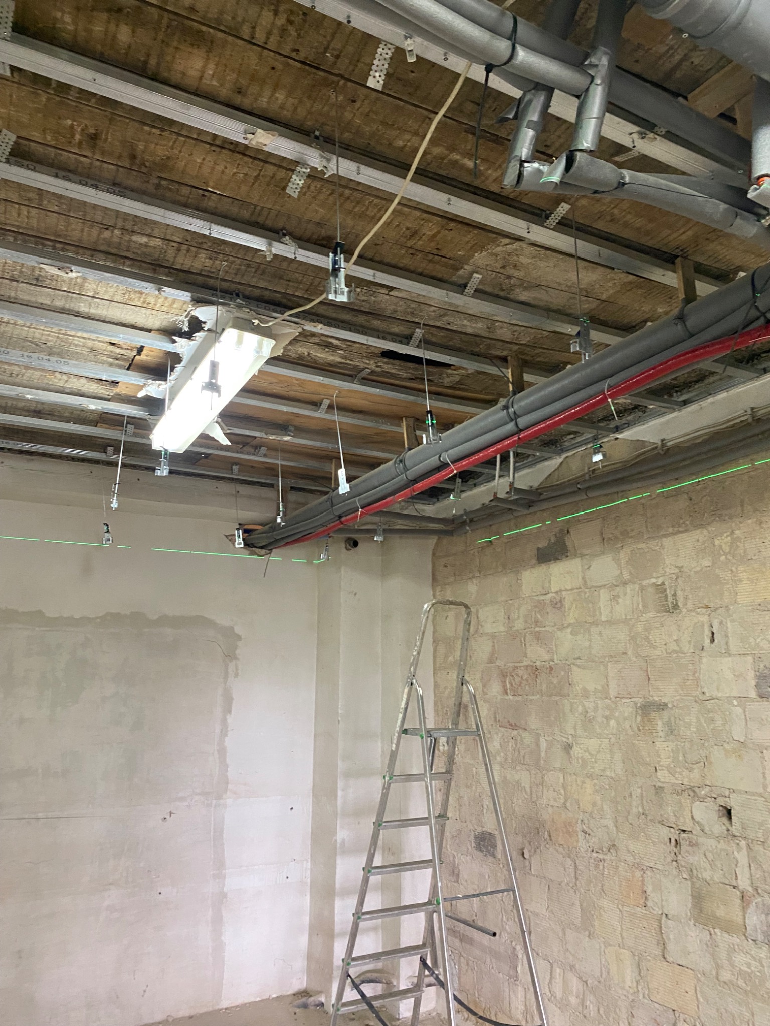 Wnętrze w trakcie remontu z widocznymi rurami instalacji grzewczej i hydraulicznej pod sufitem, oznaczonymi poziomnicą laserową, drabina oparta o surową ścianę z cegły.