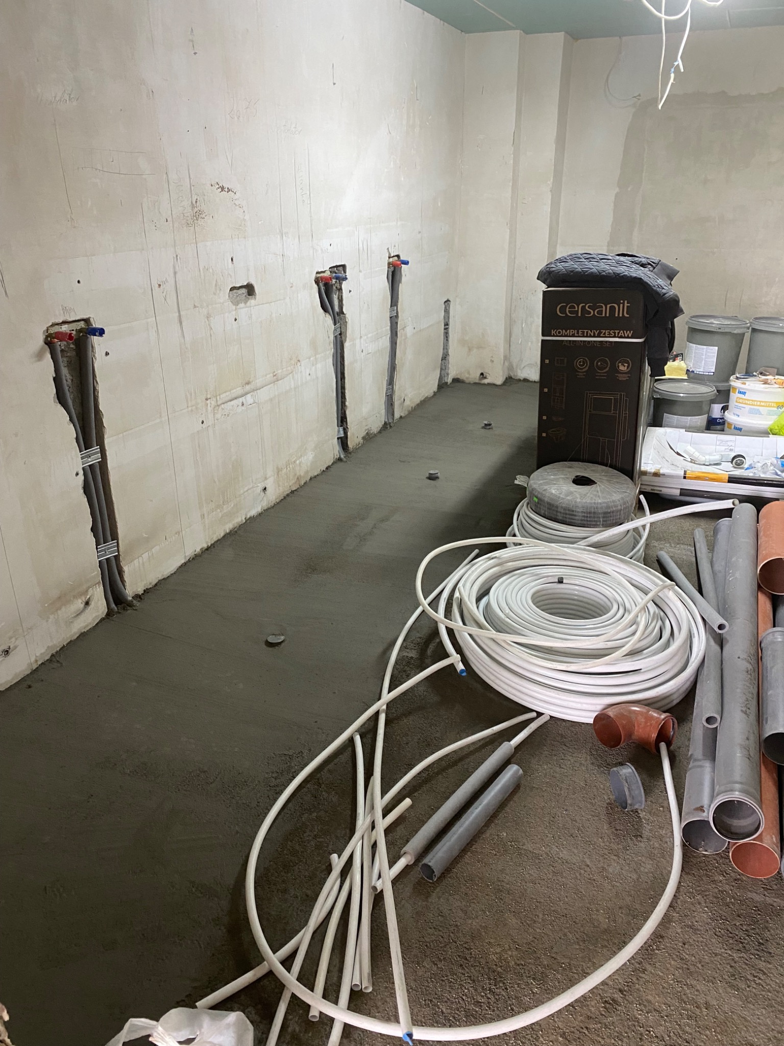 Przygotowania do instalacji wodno-kanalizacyjnej: surowe ściany z wyprowadzonymi rurami, wylewka na podłodze, zwój białych rur, szare rury kanalizacyjne i karton z zestawem Cersanit.