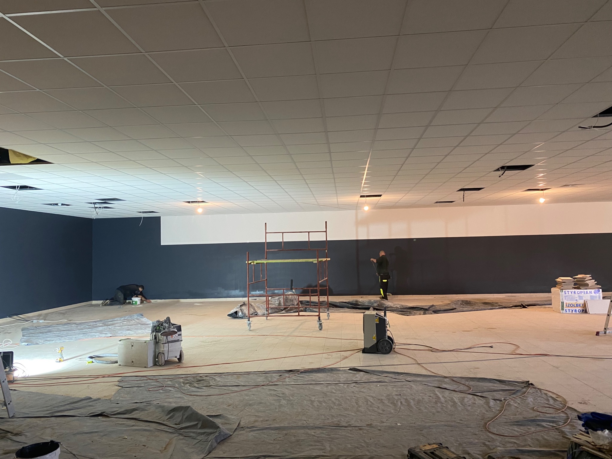 Malowanie ścian w nowym lokalu: dwóch pracowników, rusztowanie, narzędzia malarskie i zabezpieczenia podłogi przed zabrudzeniem farbą.