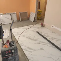 Układanie białych płytek imitujących marmur, z widocznym laserowym niwelatorem, poziomnicą i odkurzaczem przemysłowym Starmix, w trakcie prac wykończeniowych.