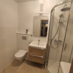 Easy Remont Rafał Lelukiewicz - Nowoczesna, jasna łazienka z kabiną prysznicową, umywalką z szafką w kolorze drewna i toaletą. W lustrze odbija się osoba sprzątająca kuchnię.
