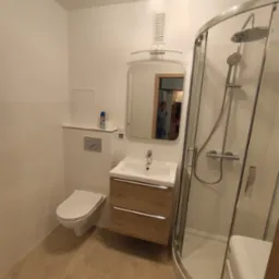 Nowoczesna, jasna łazienka z kabiną prysznicową, umywalką z szafką w kolorze drewna i toaletą. W lustrze odbija się osoba sprzątająca kuchnię.