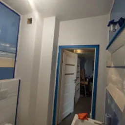 Przygotowania do malowania w łazience: ściany i framugi zabezpieczone taśmą malarską, widoczny zlew z gąbką i rękawicą, w tle otwarte drzwi z widocznym pomieszczeniem i sztalugą malarską.