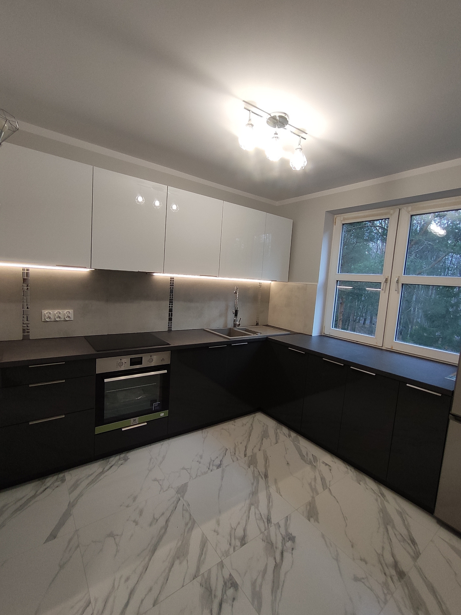 Nowoczesna kuchnia z białymi szafkami górnymi z połyskiem i czarnymi szafkami dolnymi, marmurowa podłoga, oświetlenie LED pod szafkami, widok na las za oknem.