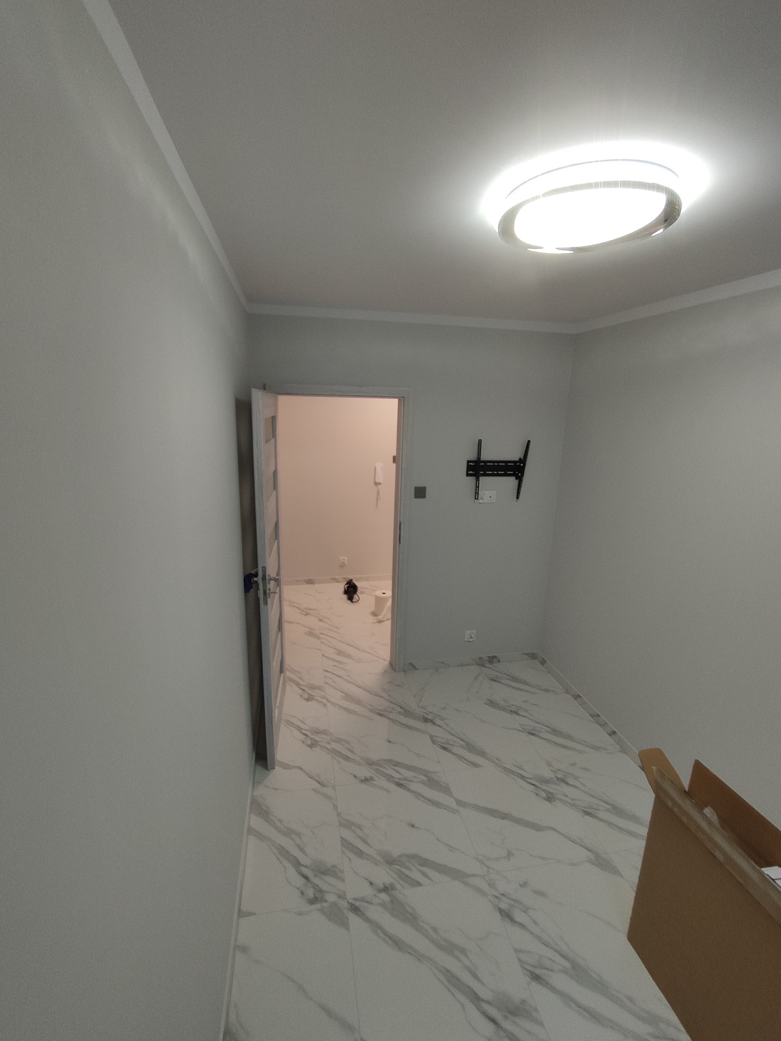 Wykończone wnętrze przedpokoju z marmurową podłogą, świeżo pomalowanymi szarymi ścianami, nowoczesną lampą sufitową i zamontowanym uchwytem na telewizor. W tle widoczne otwarte drzwi do łazienki...