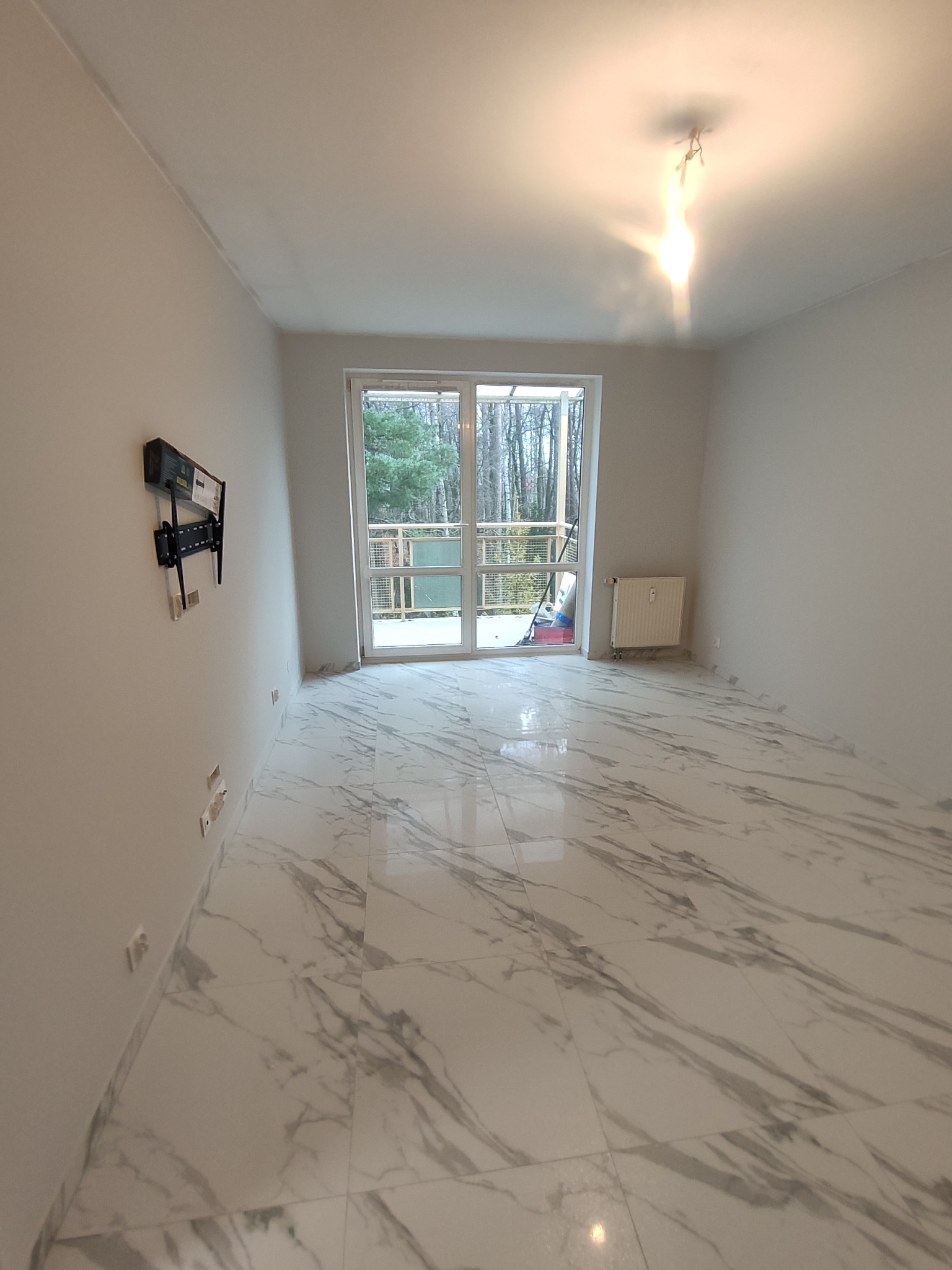 Wnętrze pokoju po remoncie z jasnymi ścianami, podłogą z płytek imitujących marmur, zamontowanym uchwytem na telewizor na ścianie i widokiem na zewnątrz przez balkonowe okno.