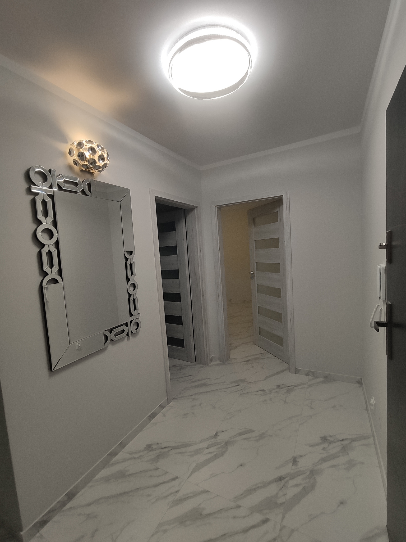 Wykończony korytarz z marmurową posadzką, ozdobnym lustrem z kryształkami i nowoczesnym oświetleniem sufitowym.