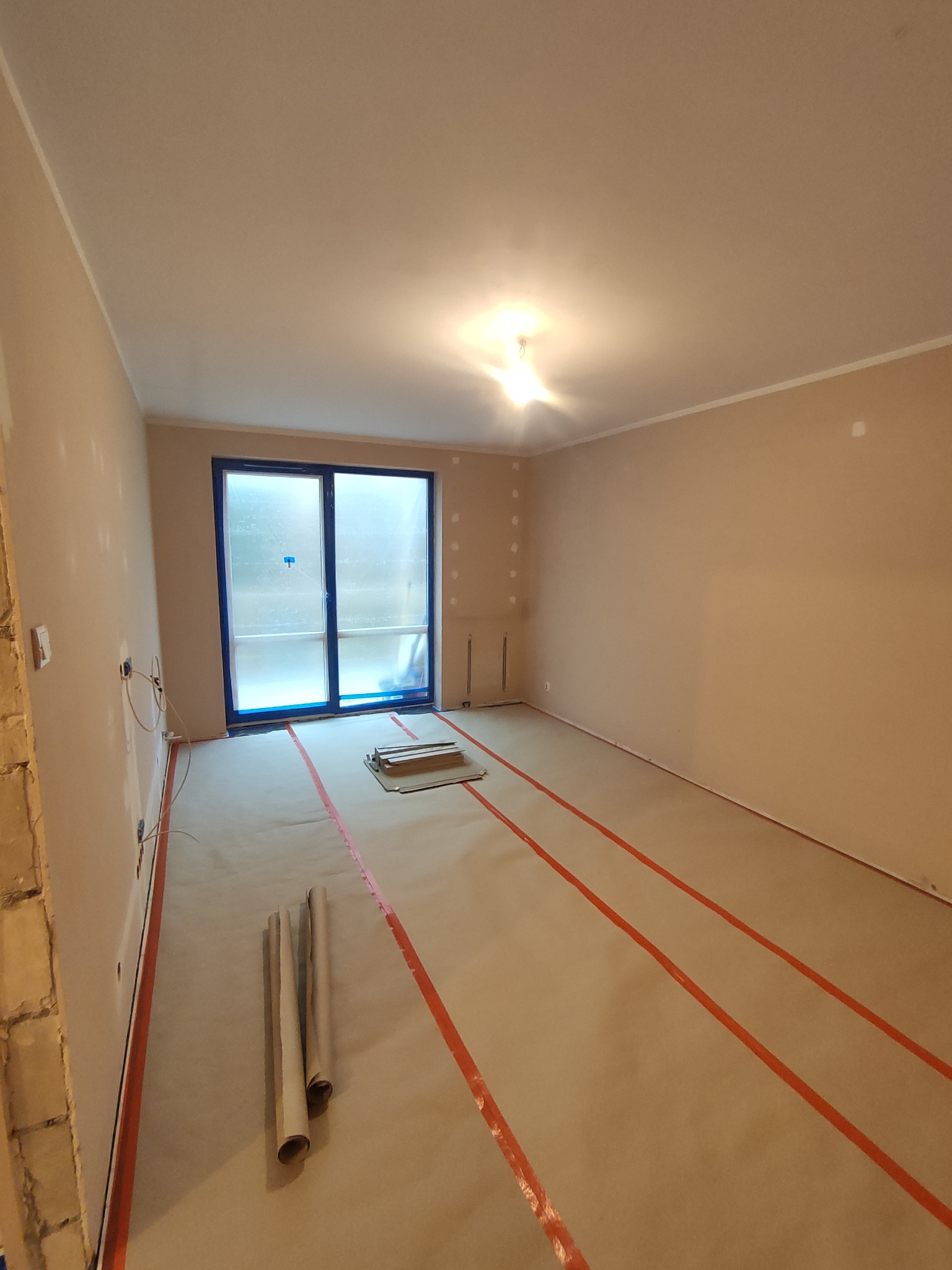 Przygotowane pomieszczenie do prac wykończeniowych: zabezpieczona podłoga papierem i taśmą, ściany wstępnie przygotowane pod malowanie lub tapetowanie, okno zabezpieczone folią.