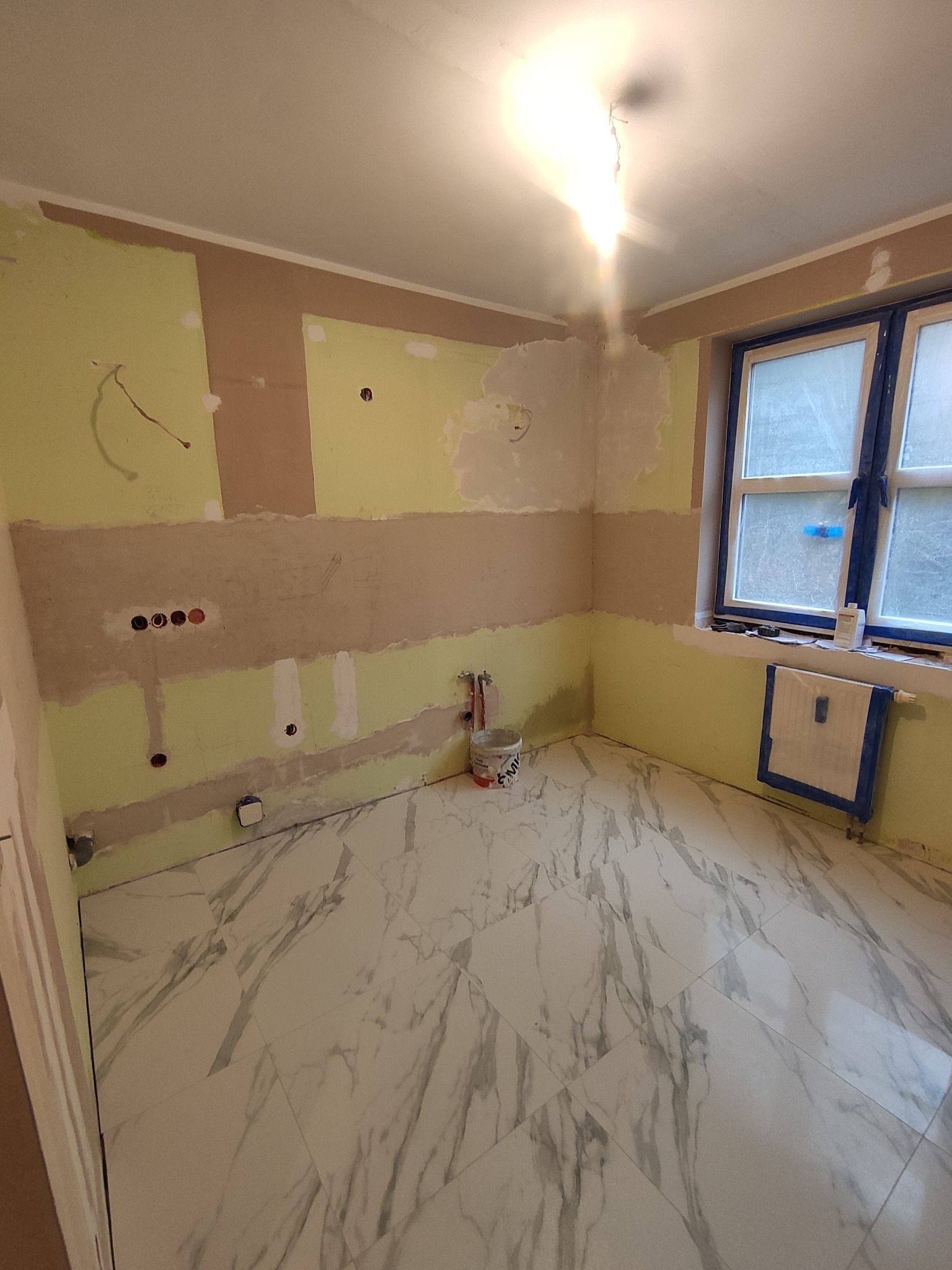 Wnętrze łazienki w trakcie prac: ściany częściowo przygotowane pod malowanie, widoczne instalacje elektryczne i hydrauliczne, na podłodze białe płytki imitujące marmur, okno zabezpieczone taśmą...