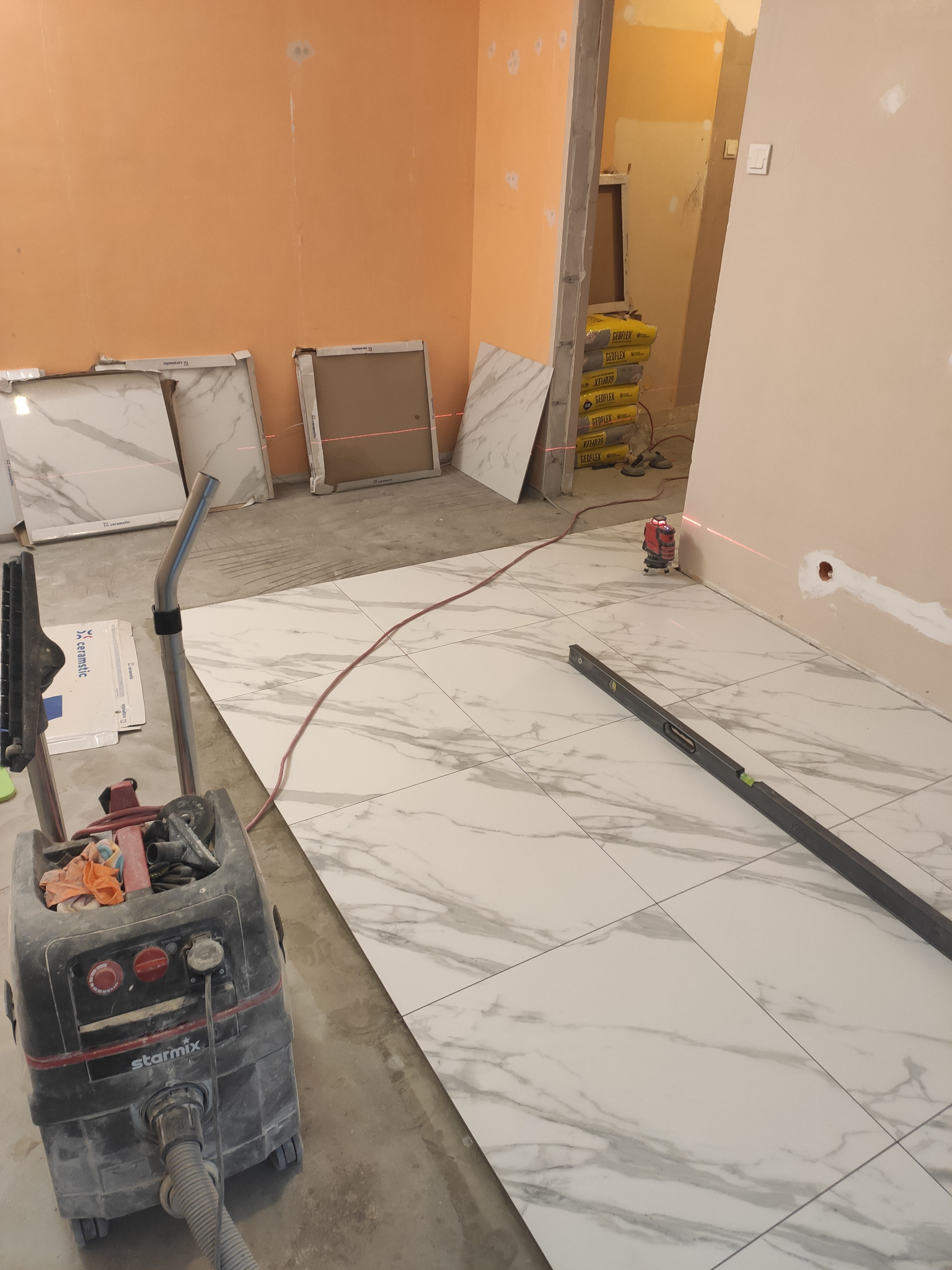 Układanie białych płytek imitujących marmur, z widocznym laserowym niwelatorem, poziomnicą i odkurzaczem przemysłowym Starmix, w trakcie prac wykończeniowych.