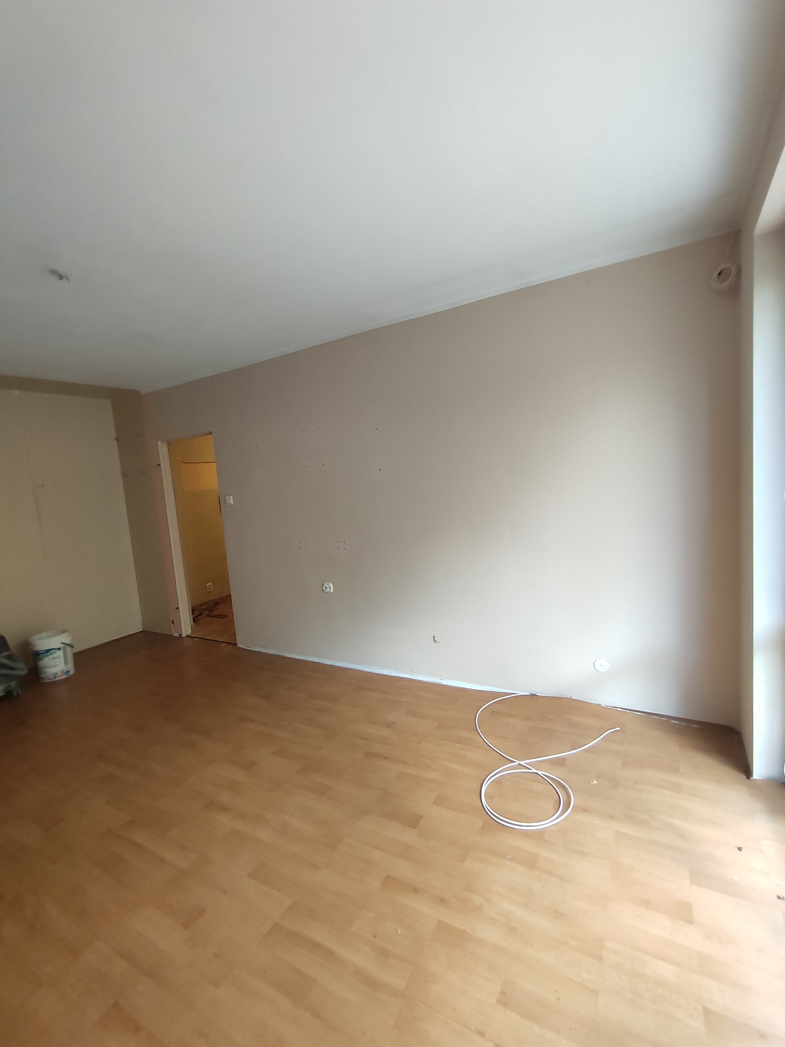 Pusty pokój przed remontem: ściany w odcieniach beżu z widocznymi śladami po kołkach, drewniana podłoga, zwój kabla elektrycznego na podłodze, widoczne wiadro z farbą w rogu pokoju.