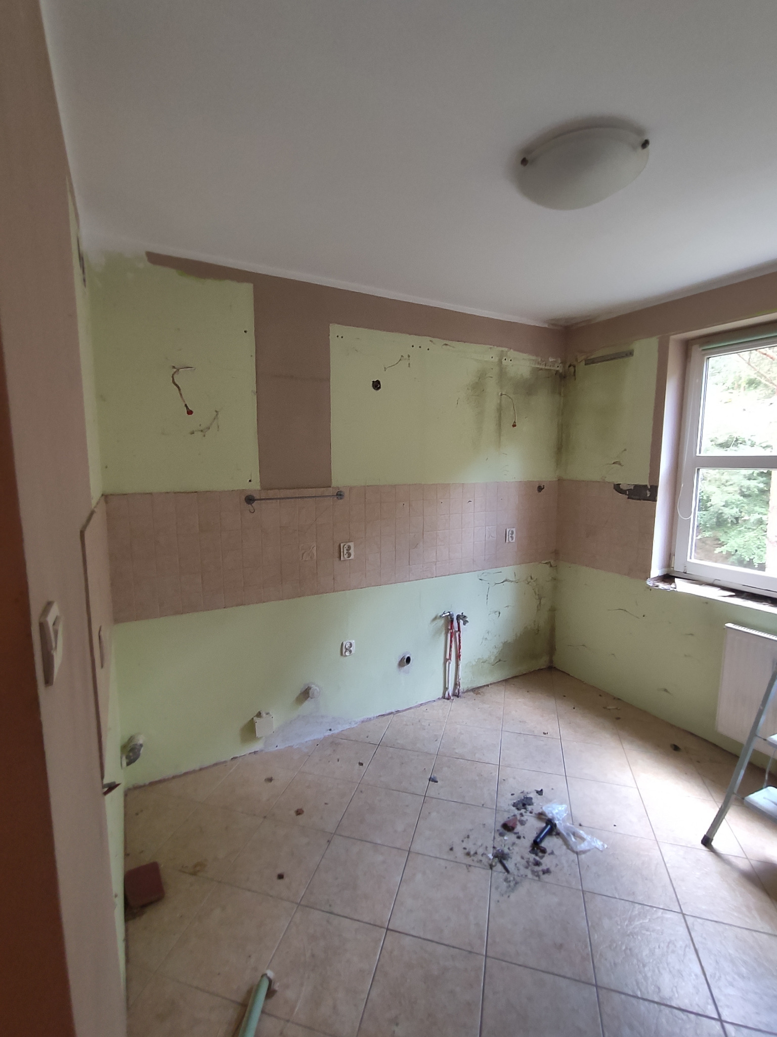 Kuchnia w trakcie demontażu, widoczne ślady po starych płytkach, odsłonięte instalacje, resztki gruzu na podłodze i drabina oparta o ścianę.