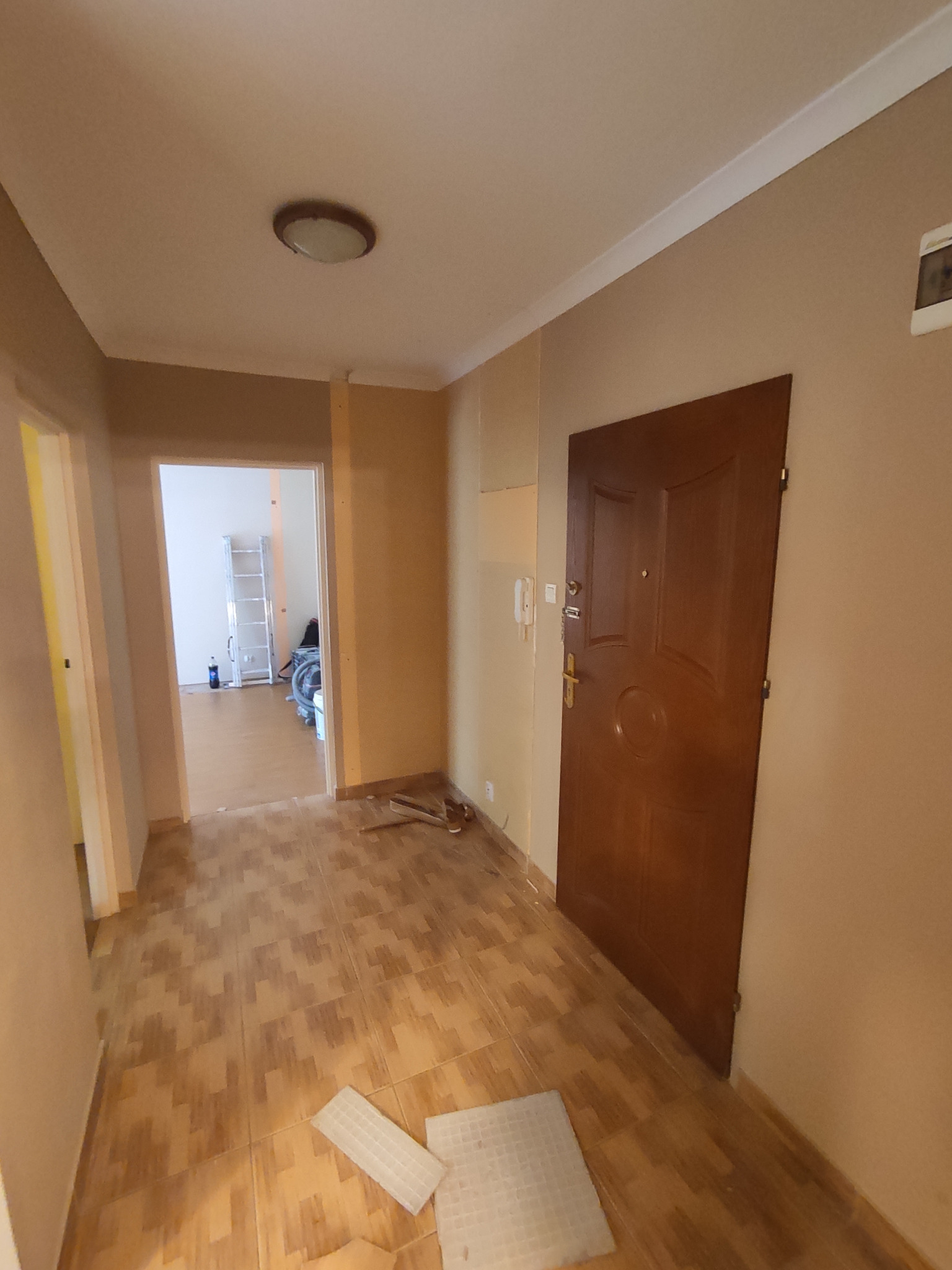 Korytarz w trakcie prac remontowych: ułożona podłoga z płytek imitujących drewno, ściany przygotowane do malowania, widoczne narzędzia i resztki materiałów, w tle drabina i sprzęt budowlany.