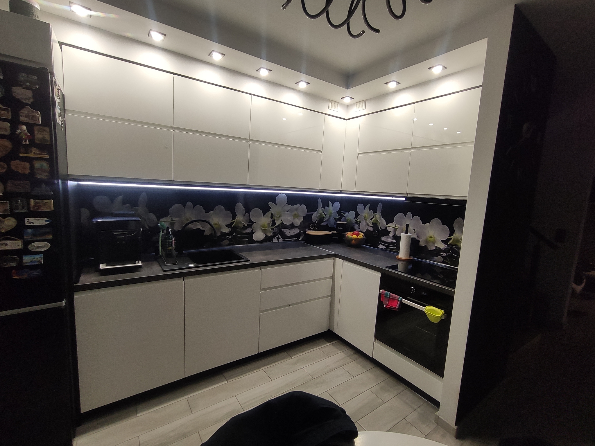 Nowoczesna kuchnia z białymi, błyszczącymi frontami szafek, czarnym blatem i panelem ściennym z motywem białych kwiatów, oświetlona punktowymi lampami sufitowymi i listwą LED pod szafkami górnymi.