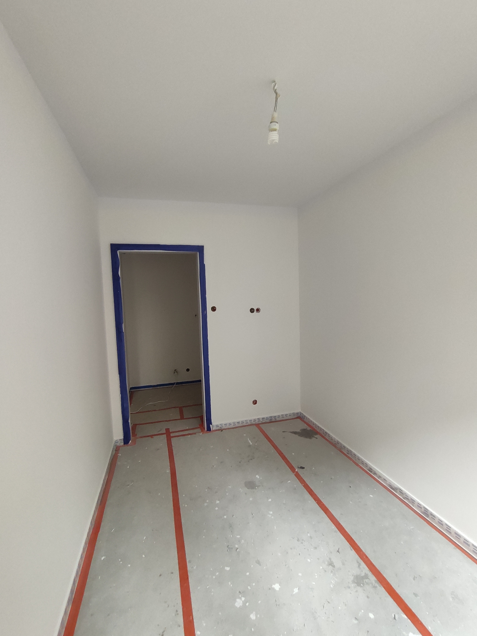 Pusty, świeżo pomalowany pokój z zabezpieczonymi listwami przypodłogowymi czerwoną taśmą, niebieska taśma malarska wokół futryny drzwiowej, surowy beton na podłodze, wystające kable z sufitu...