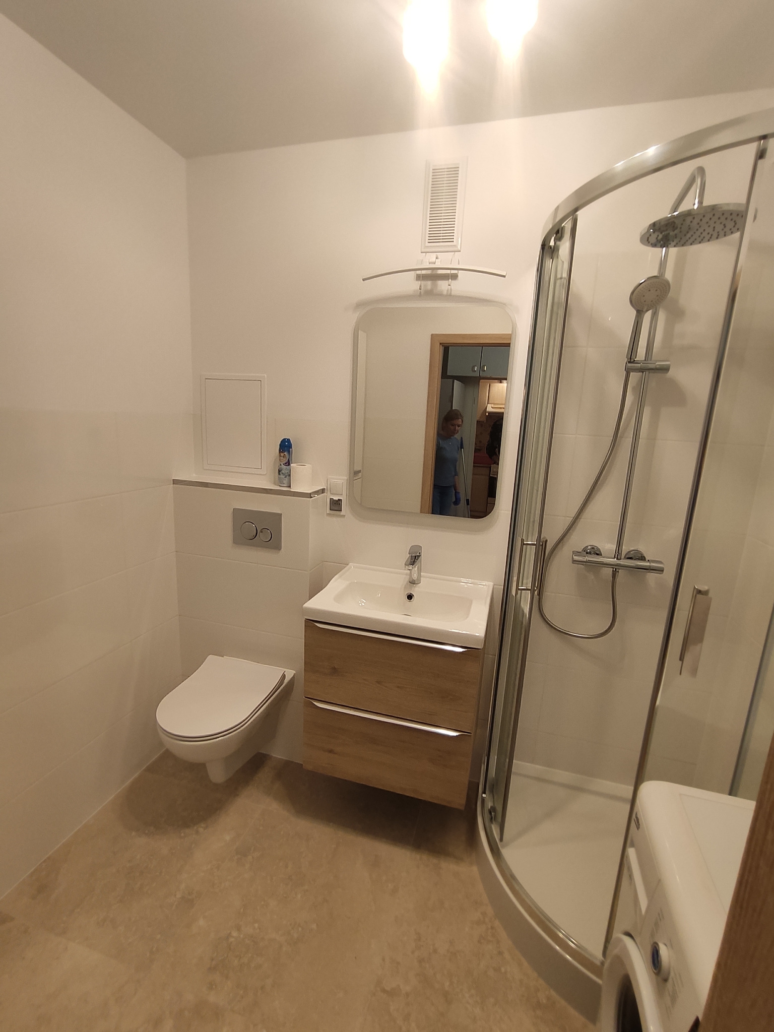 Nowoczesna, jasna łazienka z kabiną prysznicową, umywalką z szafką w kolorze drewna i toaletą. W lustrze odbija się osoba sprzątająca kuchnię.