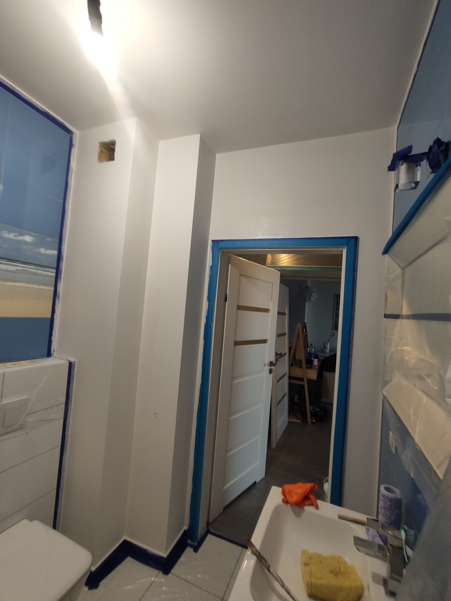 Przygotowania do malowania w łazience: ściany i framugi zabezpieczone taśmą malarską, widoczny zlew z gąbką i rękawicą, w tle otwarte drzwi z widocznym pomieszczeniem i sztalugą malarską.