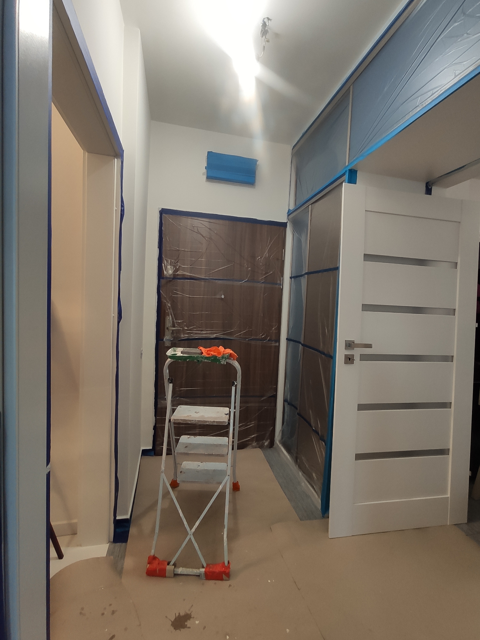 Korytarz w trakcie przygotowań do malowania: zabezpieczone folią drzwi wejściowe i szafa wnękowa, drabina malarska z pomarańczowymi rękawicami na górze, podłoga wyłożona kartonem, obrzeża ścian...