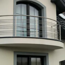 Balkon z zaokrągloną, poziomą, metalową konstrukcją zabezpieczającą, widok z zewnątrz, na tle okna z czarną ramą i fragmentu dachu z anteną.