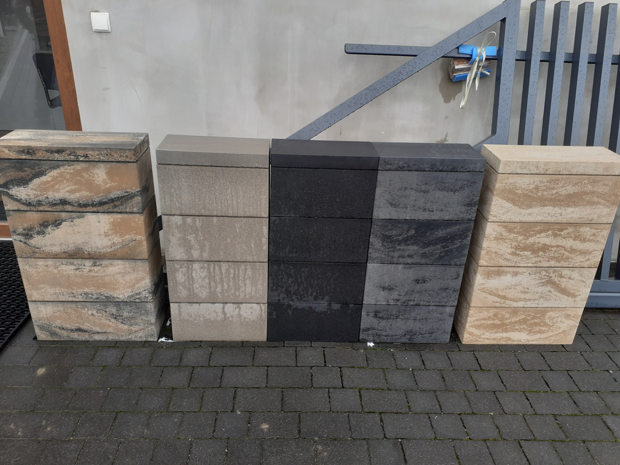 Ułożone obok siebie słupki ogrodzeniowe z betonu w różnych kolorach (beżowy, szary, czarny) na kostce brukowej, widoczne elementy ogrodzenia panelowego w tle.