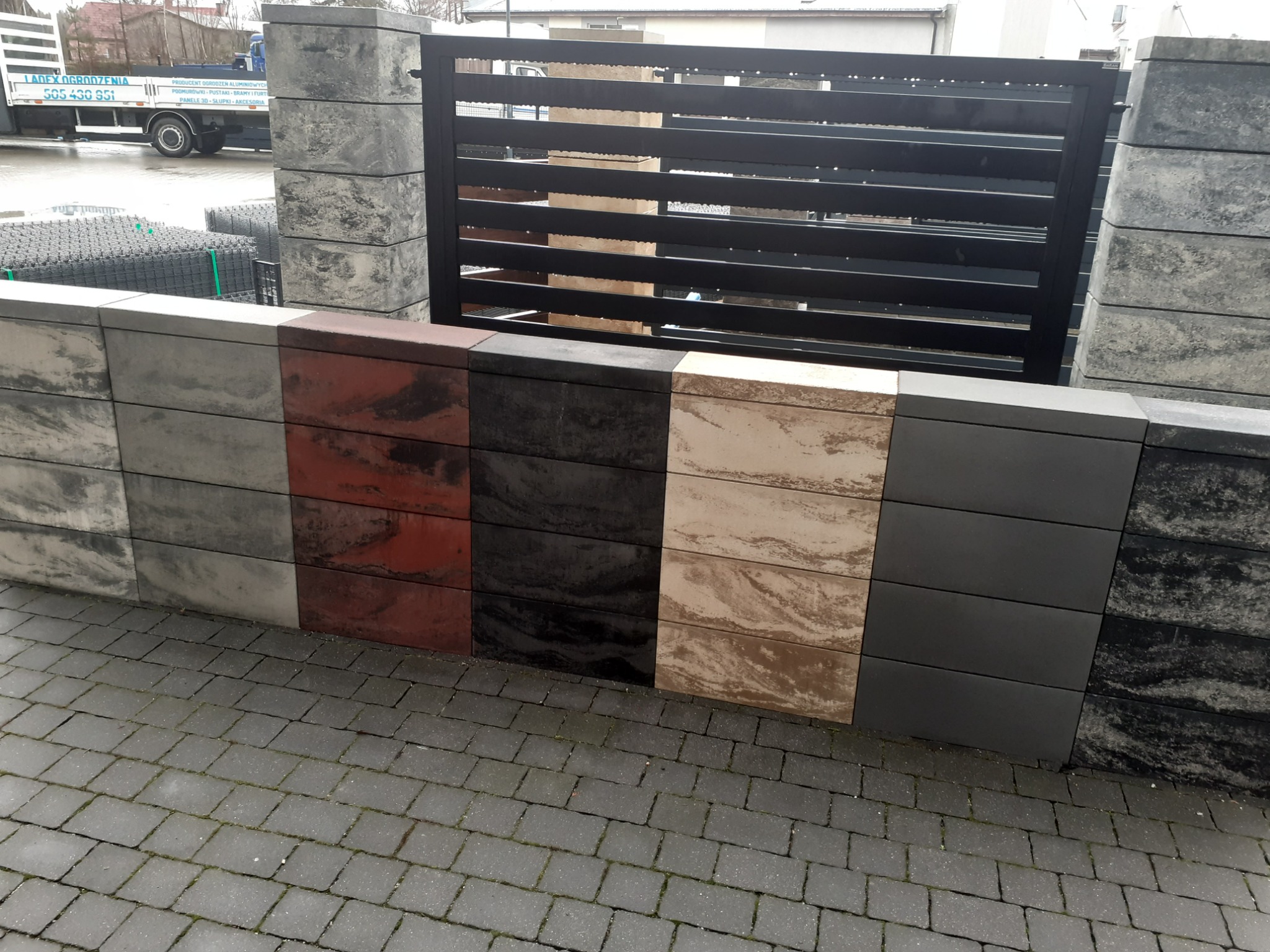 Prezentacja różnokolorowych podmurówek ogrodzeniowych z betonowych bloczków, uzupełniona o fragment metalowej bramy i kostkę brukową na ziemi, widoczne ogrodzenia panelowe w tle.