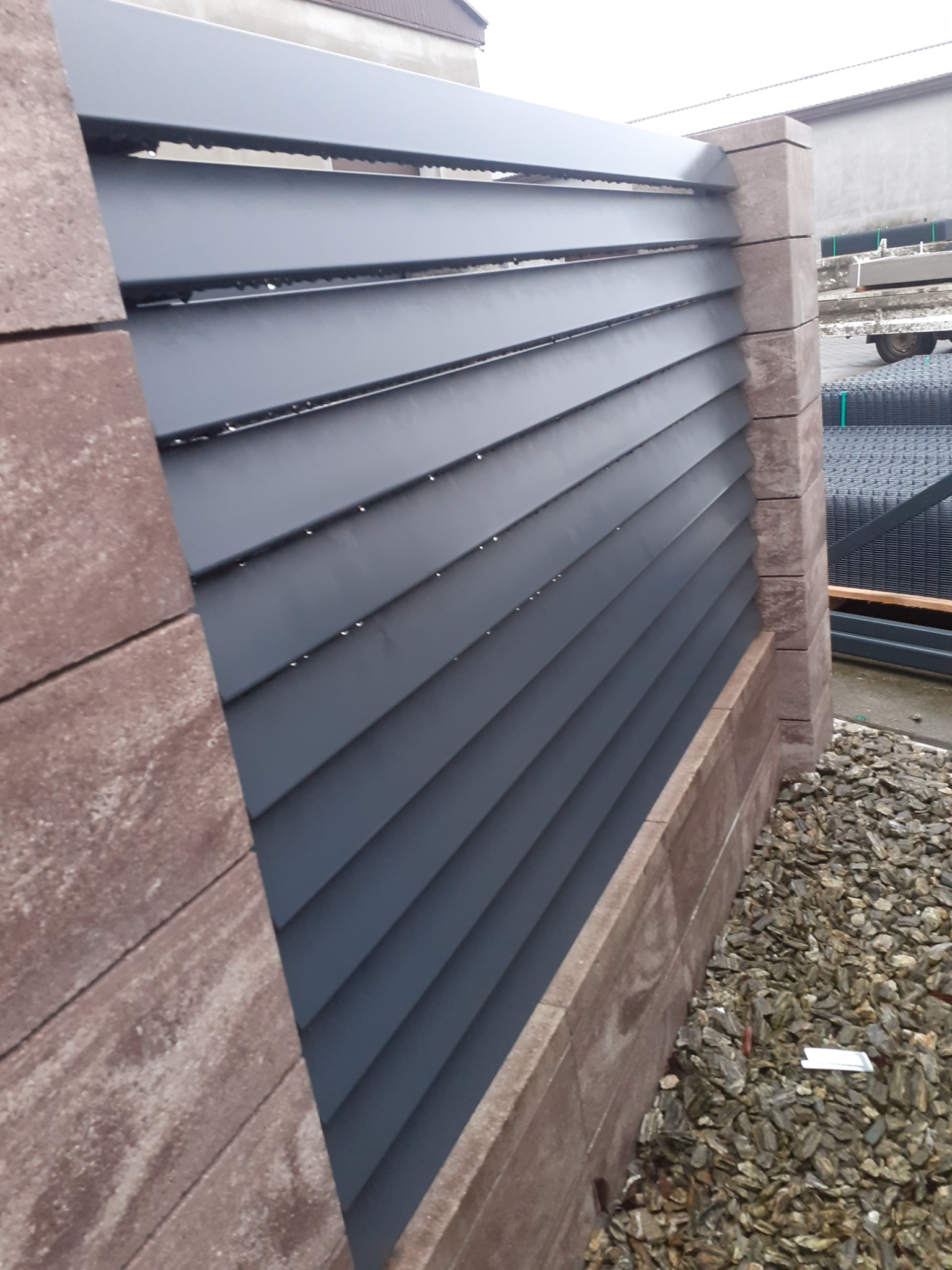 Fragment nowoczesnego ogrodzenia panelowego z poziomych, grafitowych listew, wspartego na słupkach z brązowych bloczków imitujących kamień, z widocznym podsypanym kamieniem ozdobnym u podstawy.