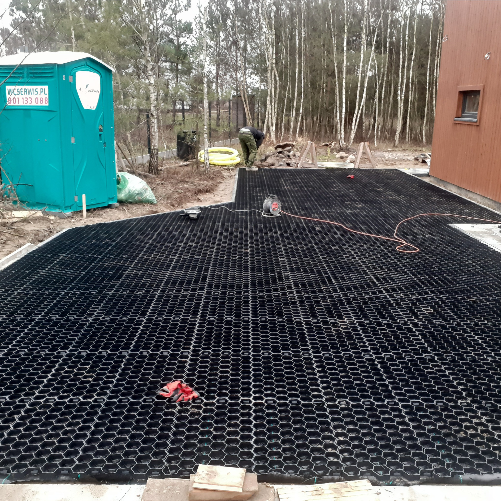 Czarny, ażurowy materiał przypominający plaster miodu, rozłożony na przygotowanym podłożu, prawdopodobnie podjazd lub parking. W tle widoczny pracownik, przenośna toaleta w kolorze turkusowym...