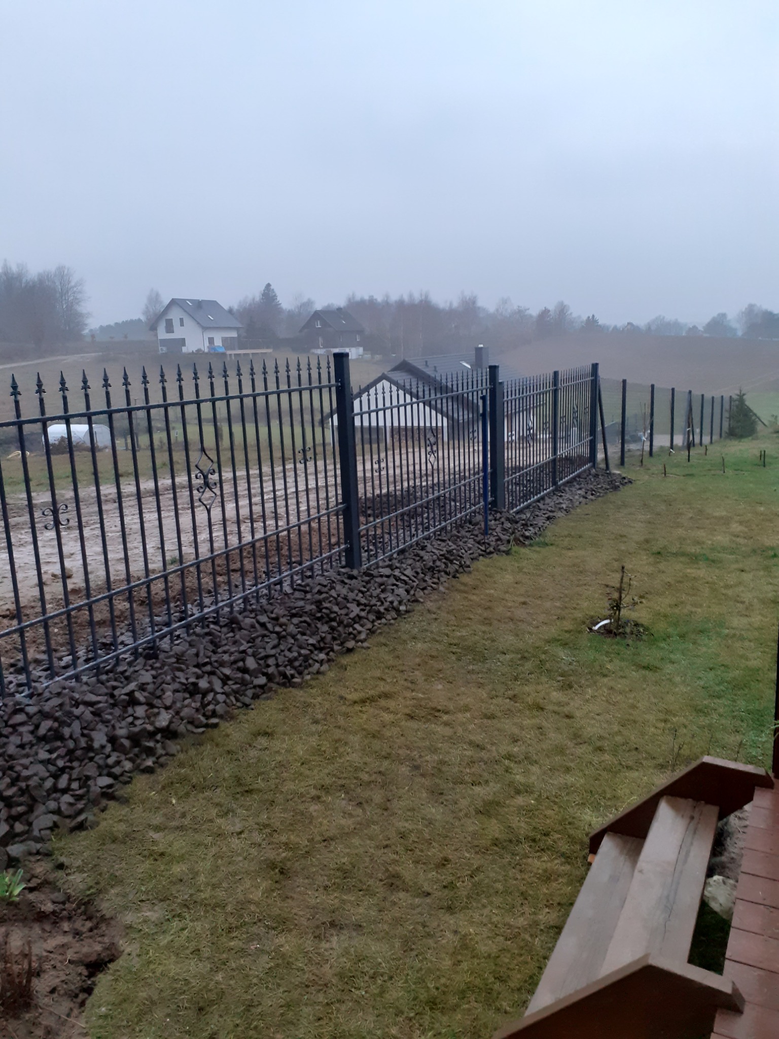 Metalowe ogrodzenie posesji z dekoracyjnymi elementami, podsypane kamieniami, widok z drewnianych schodów w pochmurny dzień.