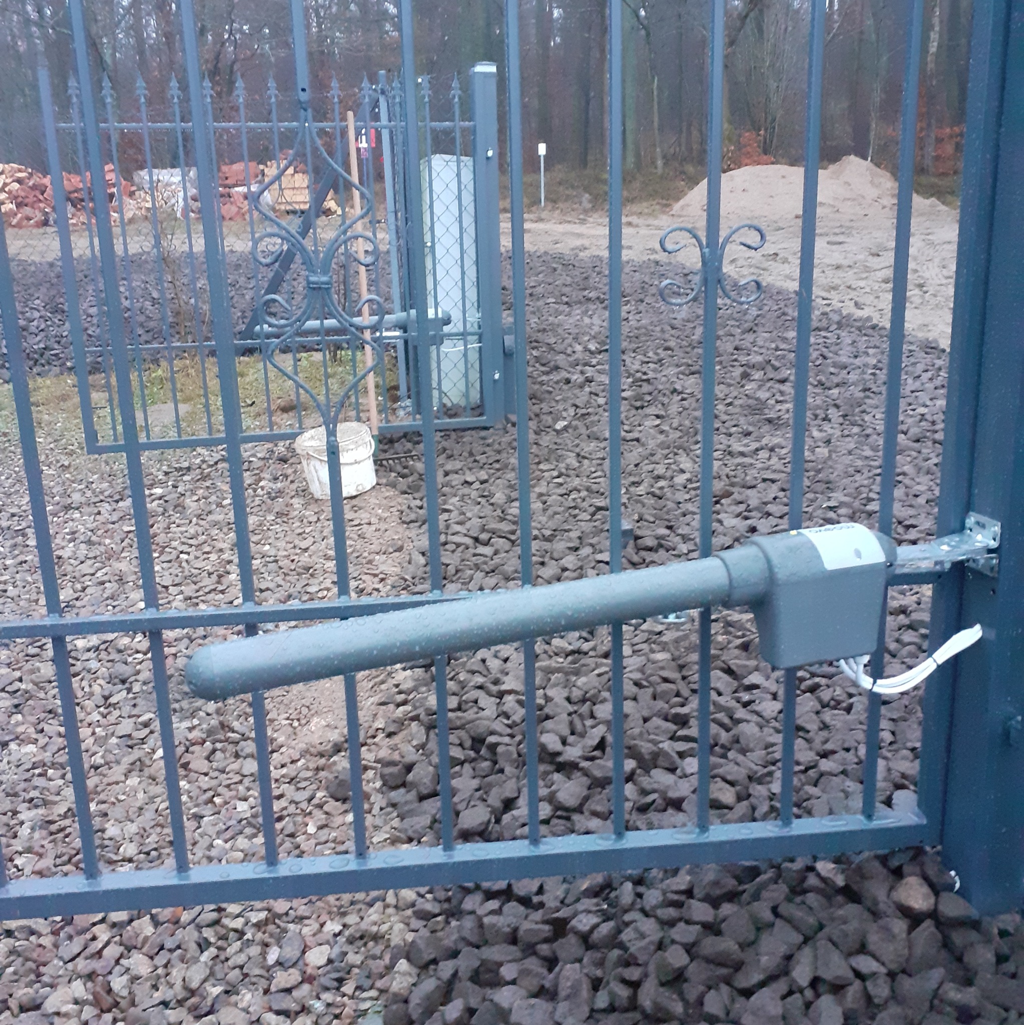 Szara metalowa brama z automatycznym napędem, widoczne elementy konstrukcyjne i mechanizm otwierający, tło z kamieniami i stosem cegieł.