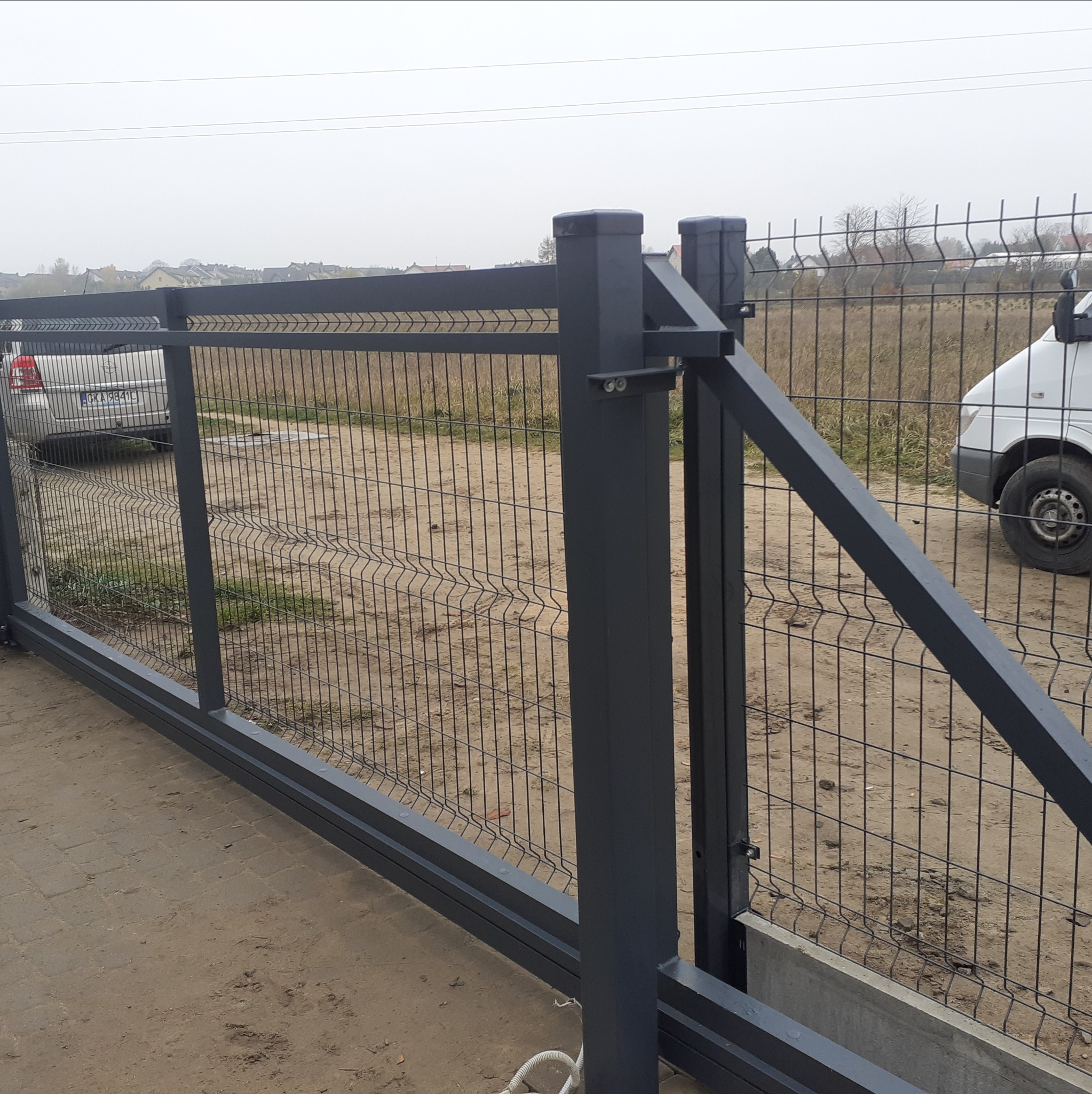 Przesuwna brama ogrodzeniowa z metalowej siatki w kolorze grafitowym, widoczny mechanizm prowadzący, w tle samochody i zabudowa jednorodzinna.