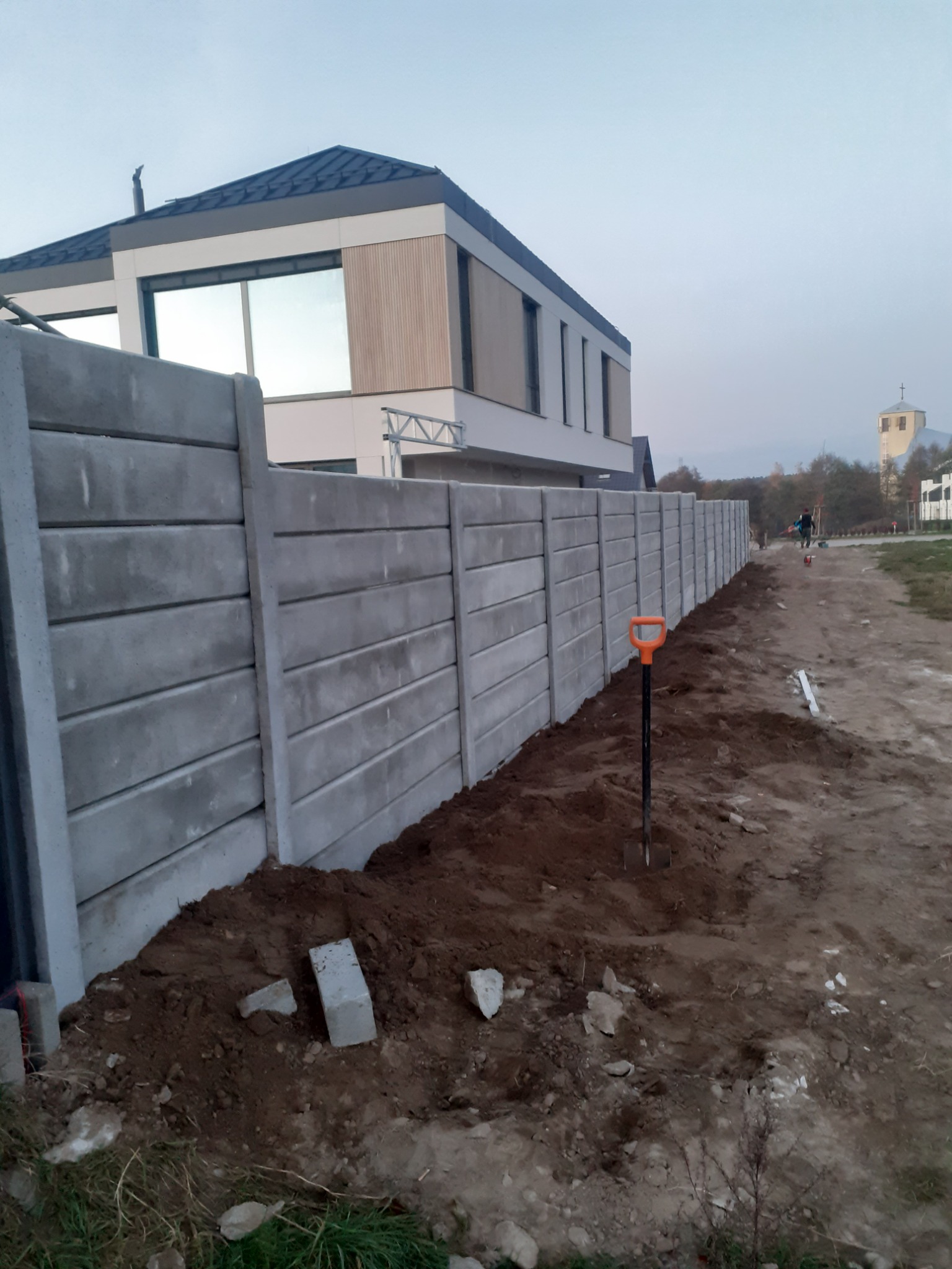 Długi, prefabrykowany, betonowy płot panelowy w trakcie montażu, widoczna pomarańczowa łopata wbita w ziemię, nowoczesny dom w tle i kościół w oddali.