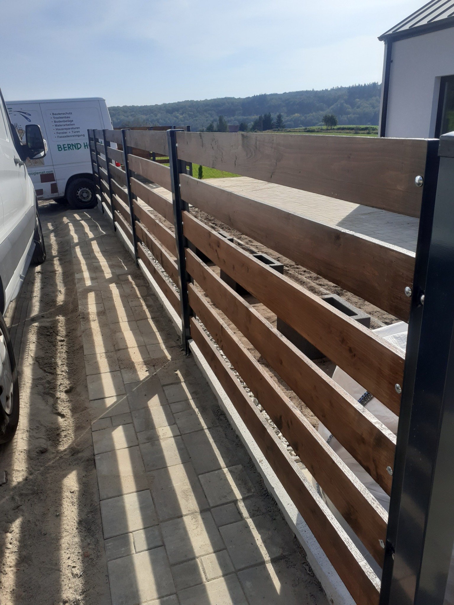 Nowoczesne ogrodzenie poziome z drewnianymi panelami i czarnymi słupkami, widoczne z boku, z częściowo ułożoną kostką brukową i samochodem dostawczym w tle.