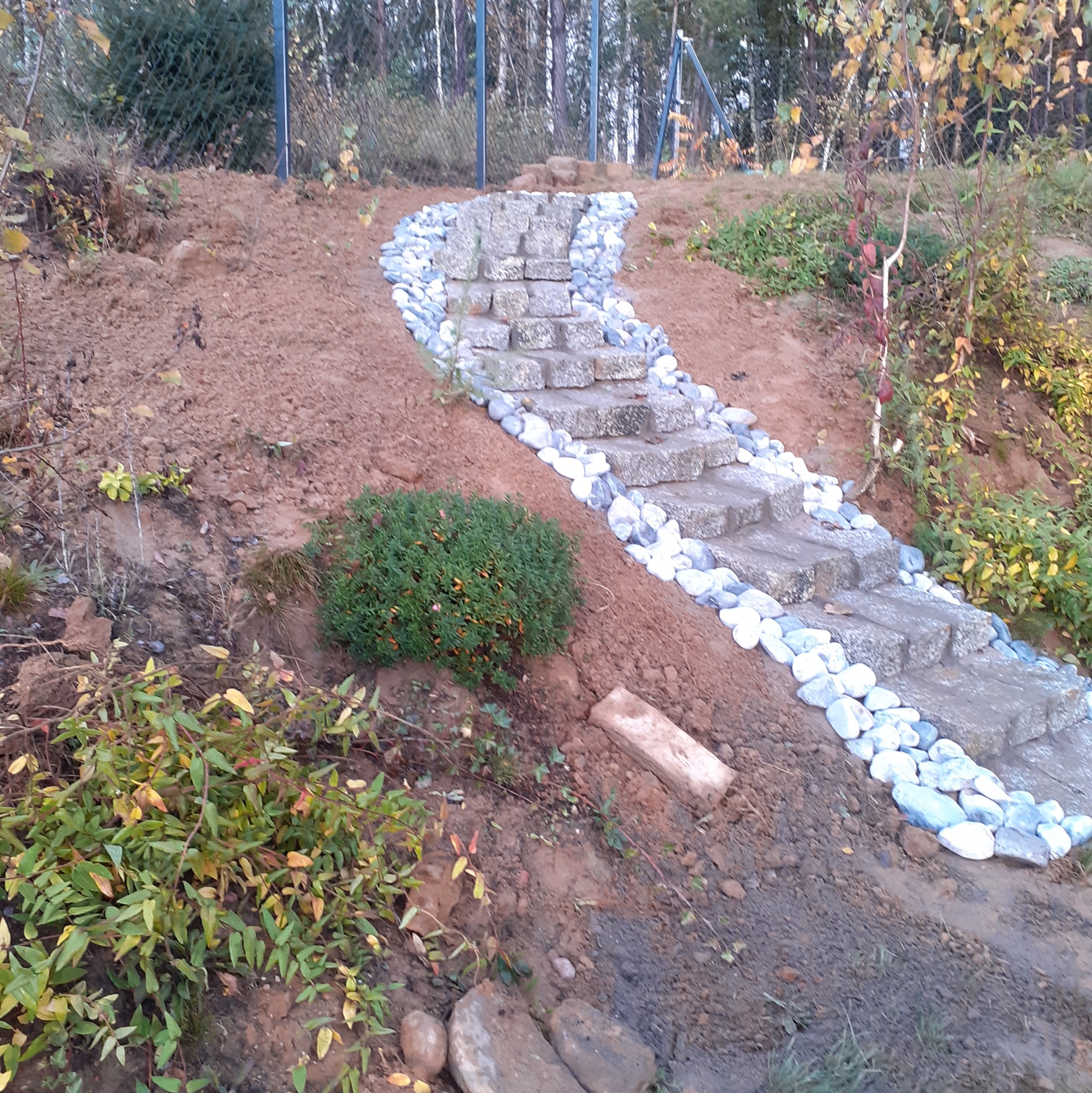 Kamienne schody ogrodowe prowadzące w górę zbocza, otoczone białymi kamieniami i roślinnością, częściowo wykończone.