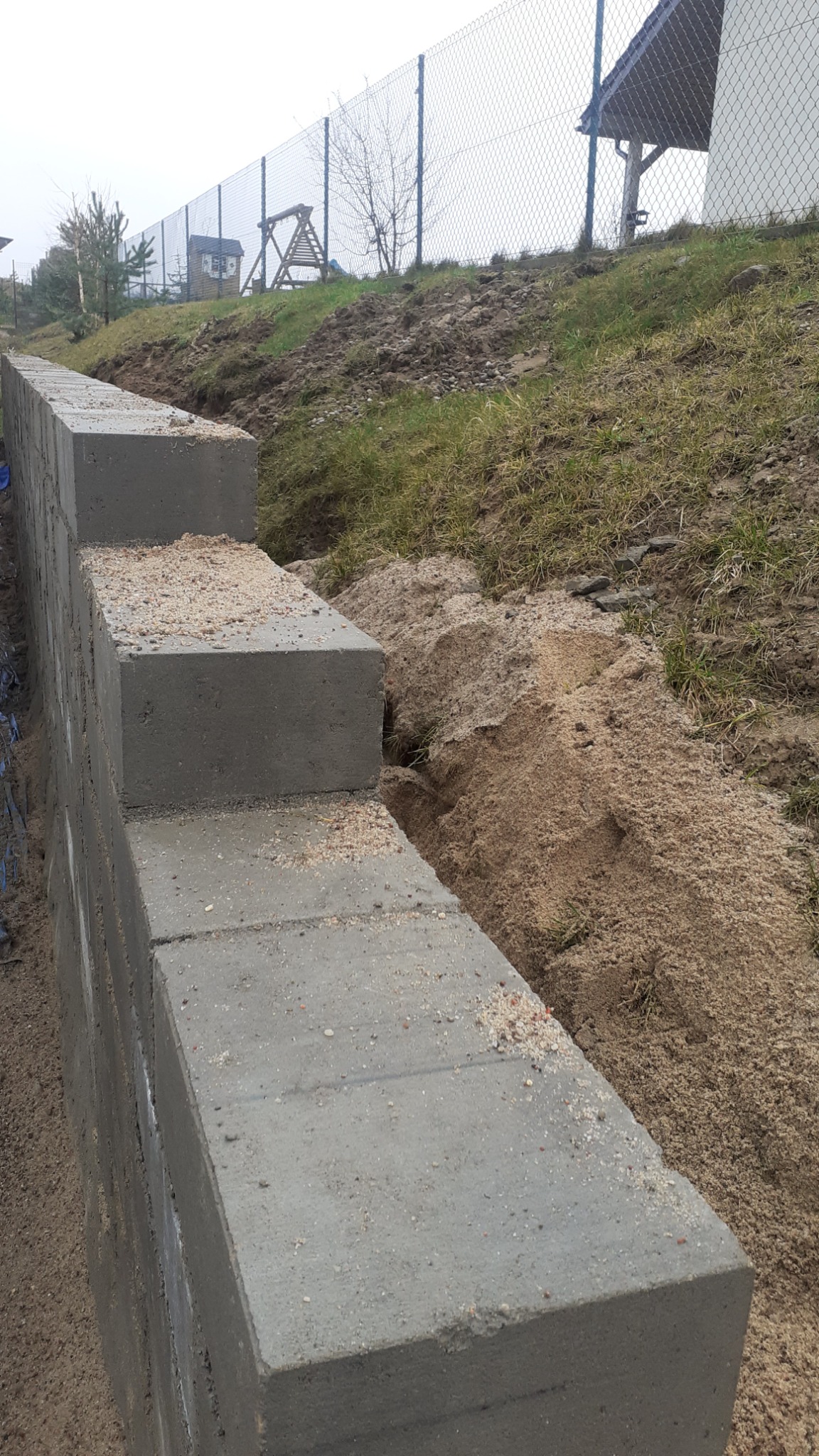 Wznoszenie muru oporowego z betonowych bloczków na nierównym terenie, widoczny wykop i ogrodzenie siatkowe w tle.