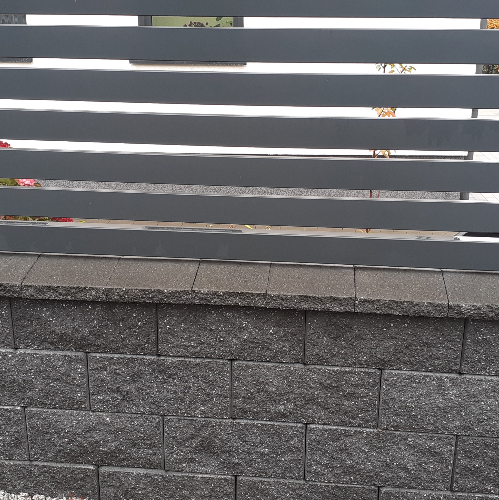 Nowoczesne ogrodzenie z poziomych, szarych paneli na murku z ciemnoszarych bloczków, widok z bliska.