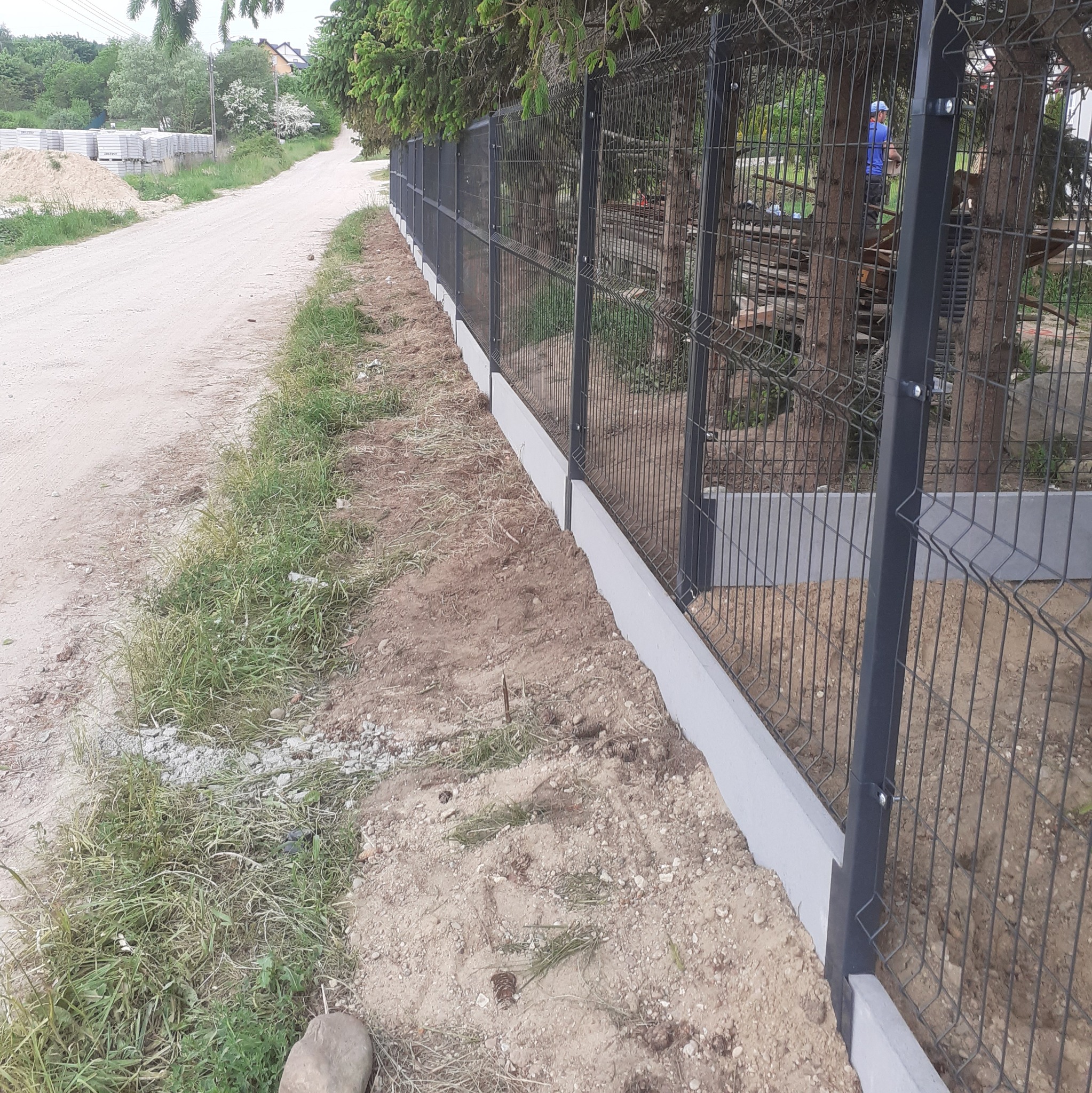 Nowo postawione, grafitowe ogrodzenie panelowe z podmurówką, widok z boku, trawa i droga szutrowa w tle.
