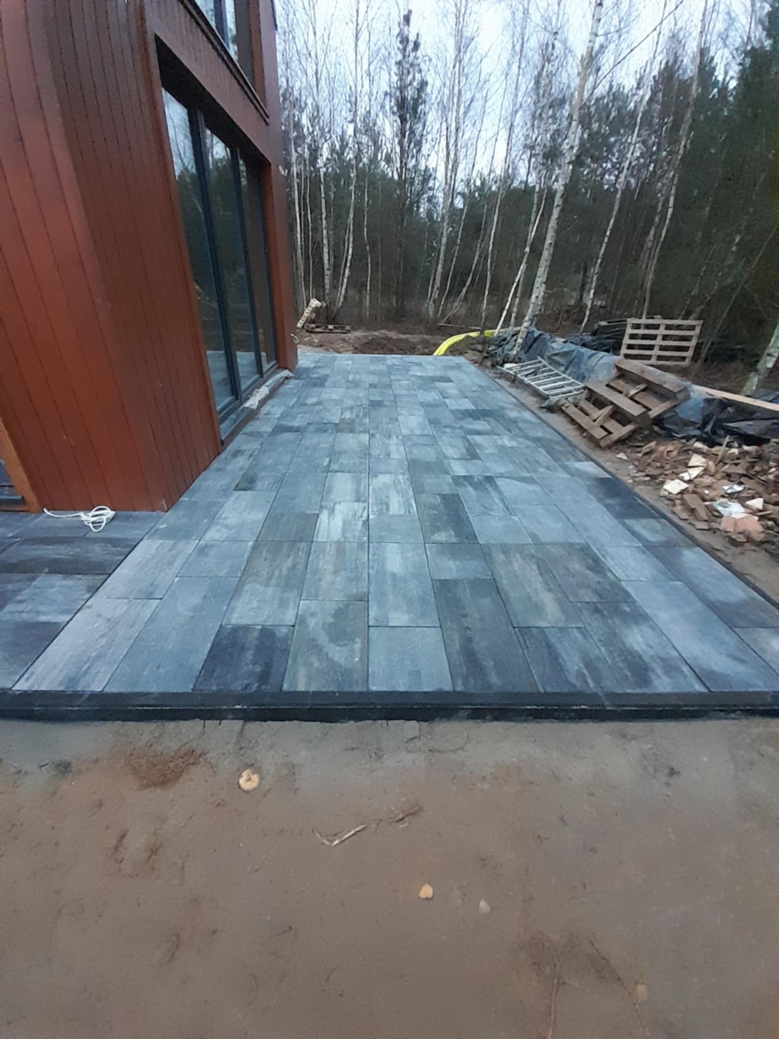 Nowo ułożona, szara kostka brukowa w geometryczny wzór, przylegająca do nowoczesnego domu z drewnianą elewacją, widok na las w tle, teren w trakcie prac wykończeniowych.