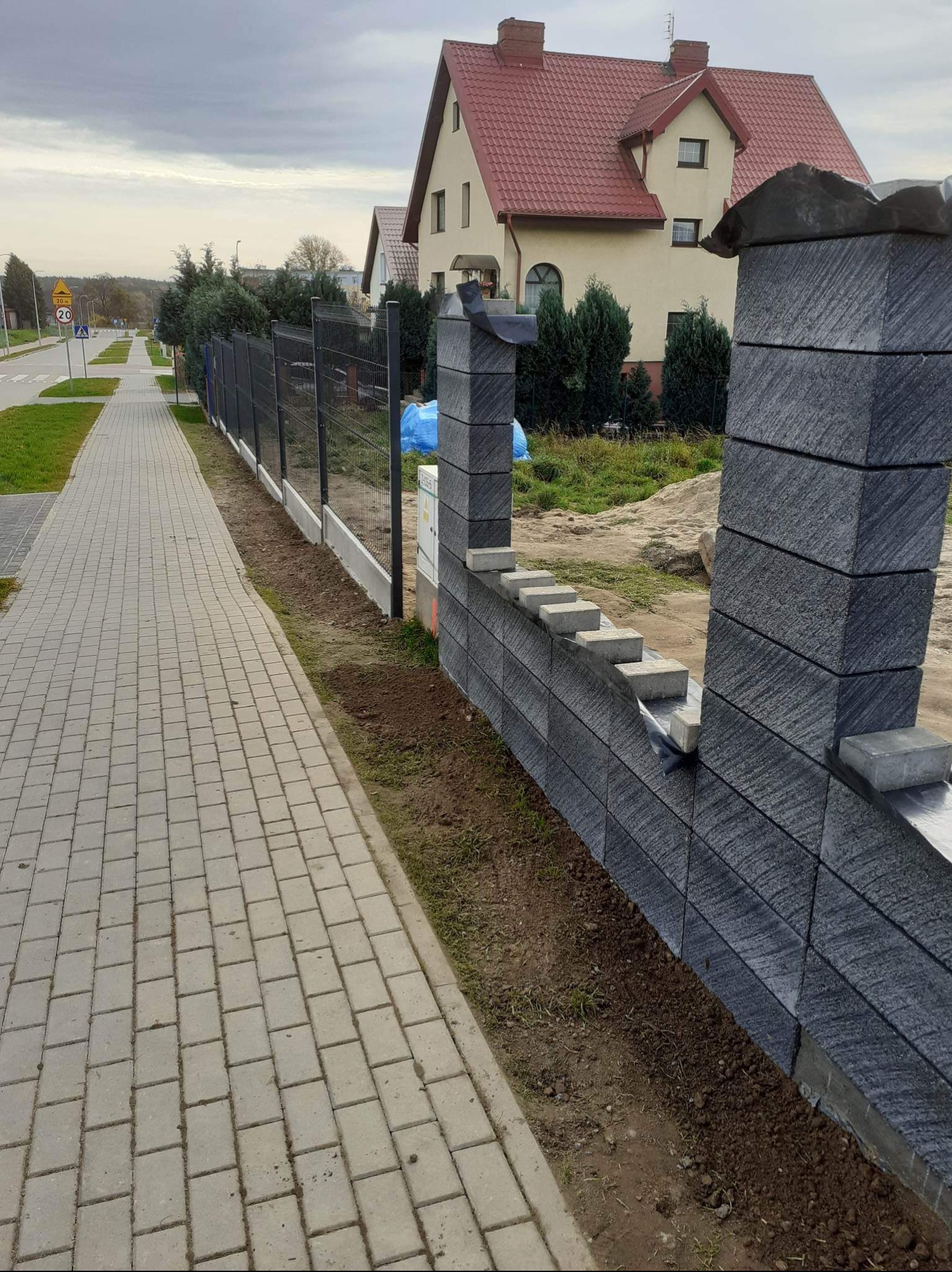 Nowoczesne ogrodzenie z szarych bloczków betonowych i metalowej siatki wzdłuż chodnika z kostki, widok na dom z czerwoną dachówką w tle.