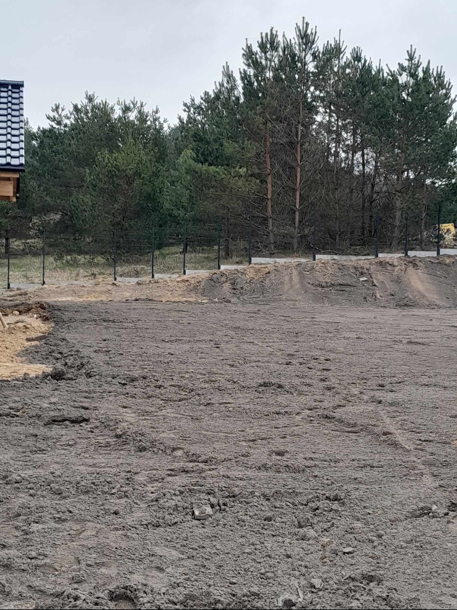 Przygotowany teren pod budowę z widocznym fragmentem fundamentu domu z czarnym ogrodzeniem siatkowym w tle i drzewami iglastymi.