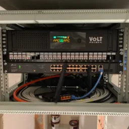 Szafa rack z zamontowanym zasilaczem awaryjnym UPS Volt Polska, switchem TP-Link oraz okablowaniem sieciowym. Widoczne porty Ethernet, diody sygnalizacyjne i kable różnej grubości.