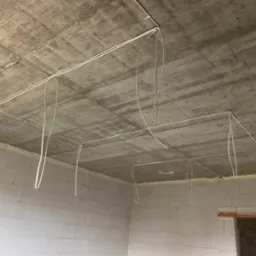 Rozprowadzone przewody elektryczne po suficie surowego pomieszczenia z białych bloczków, widoczne mocowania kabli i zwisające pętle.