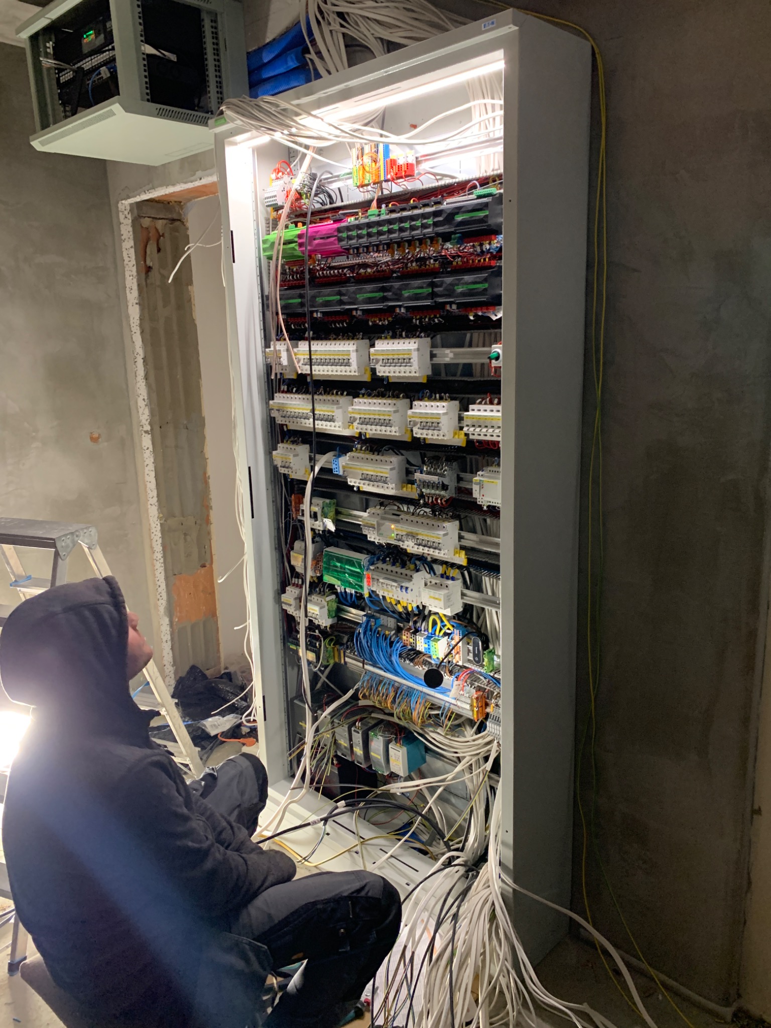 Elektryk w ciemnej bluzie siedzi przed otwartą szafą rozdzielczą, analizując skomplikowany układ przewodów, zabezpieczeń i modułów; widoczne niezabezpieczone kable zwisające na zewnątrz, surowy...