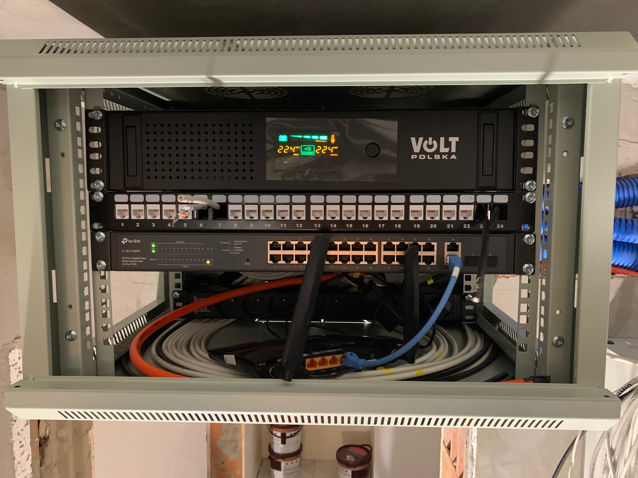 Szafa rack z zamontowanym zasilaczem awaryjnym UPS Volt Polska, switchem TP-Link oraz okablowaniem sieciowym. Widoczne porty Ethernet, diody sygnalizacyjne i kable różnej grubości.