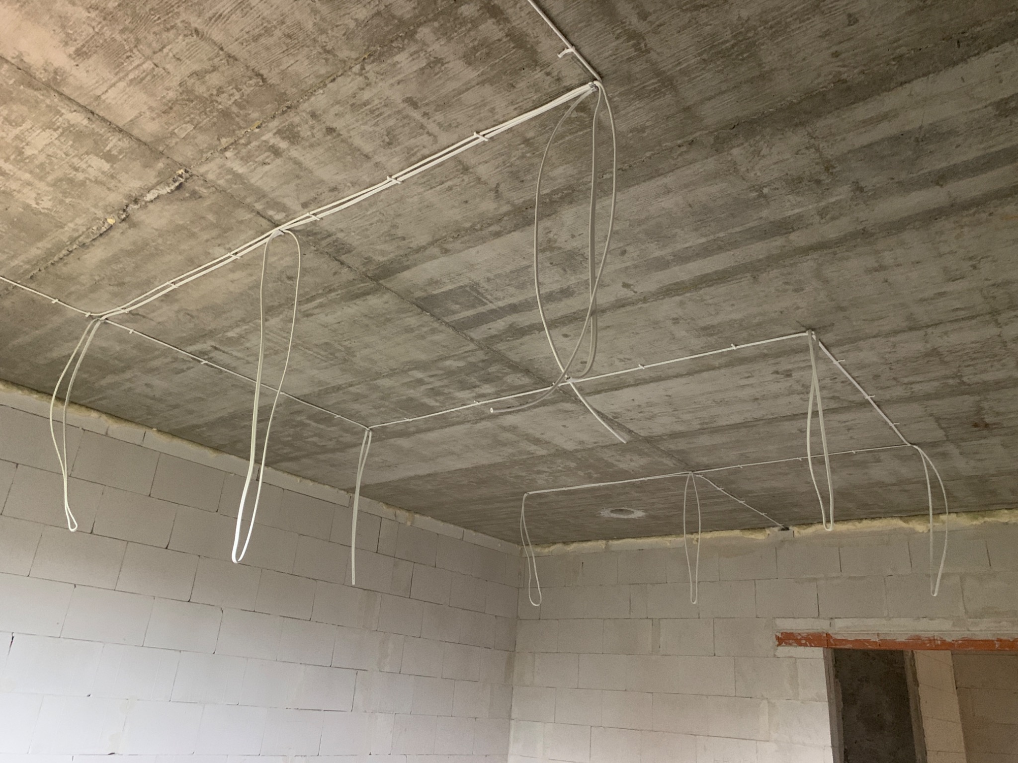 Rozprowadzone przewody elektryczne po suficie surowego pomieszczenia z białych bloczków, widoczne mocowania kabli i zwisające pętle.