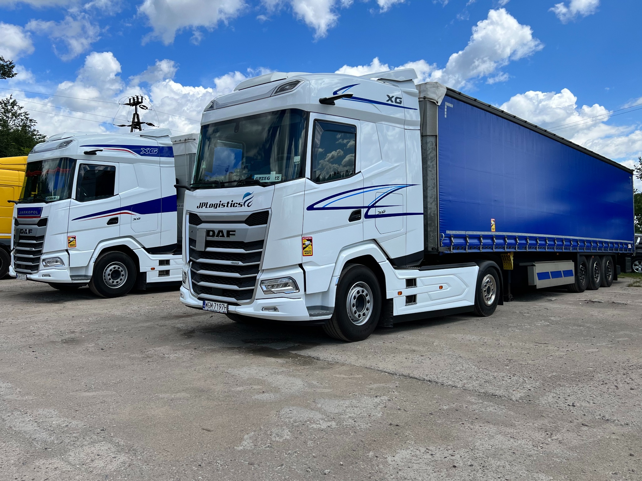 Dwa białe ciągniki siodłowe DAF XG z niebieską plandeką na naczepie, z logo JPLogistics, stojące na placu w słoneczny dzień.