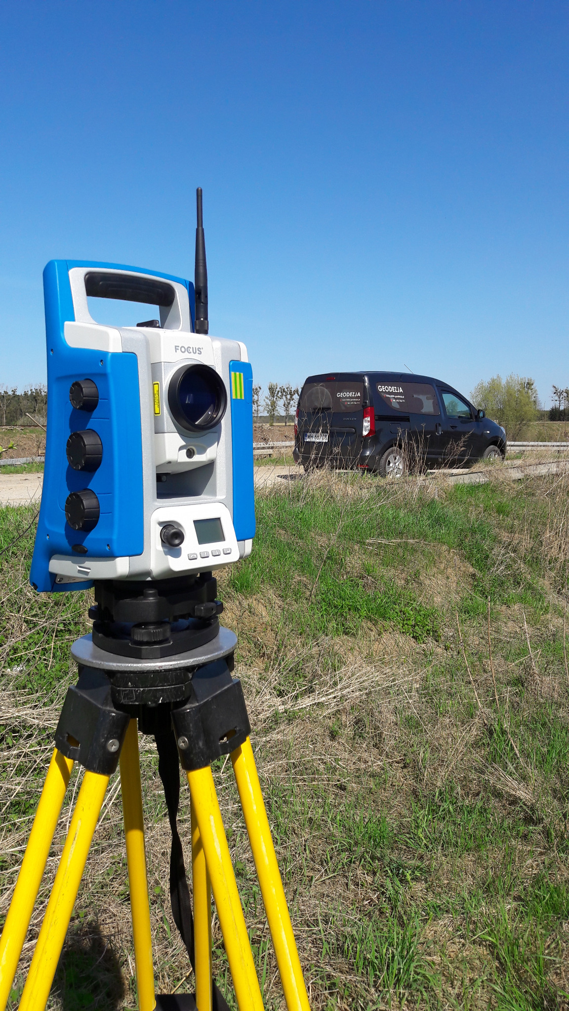 Niwelator geodezyjny Focus na żółtym statywie, w tle czarny samochód z logo firmy geodezyjnej, niebieskie niebo.