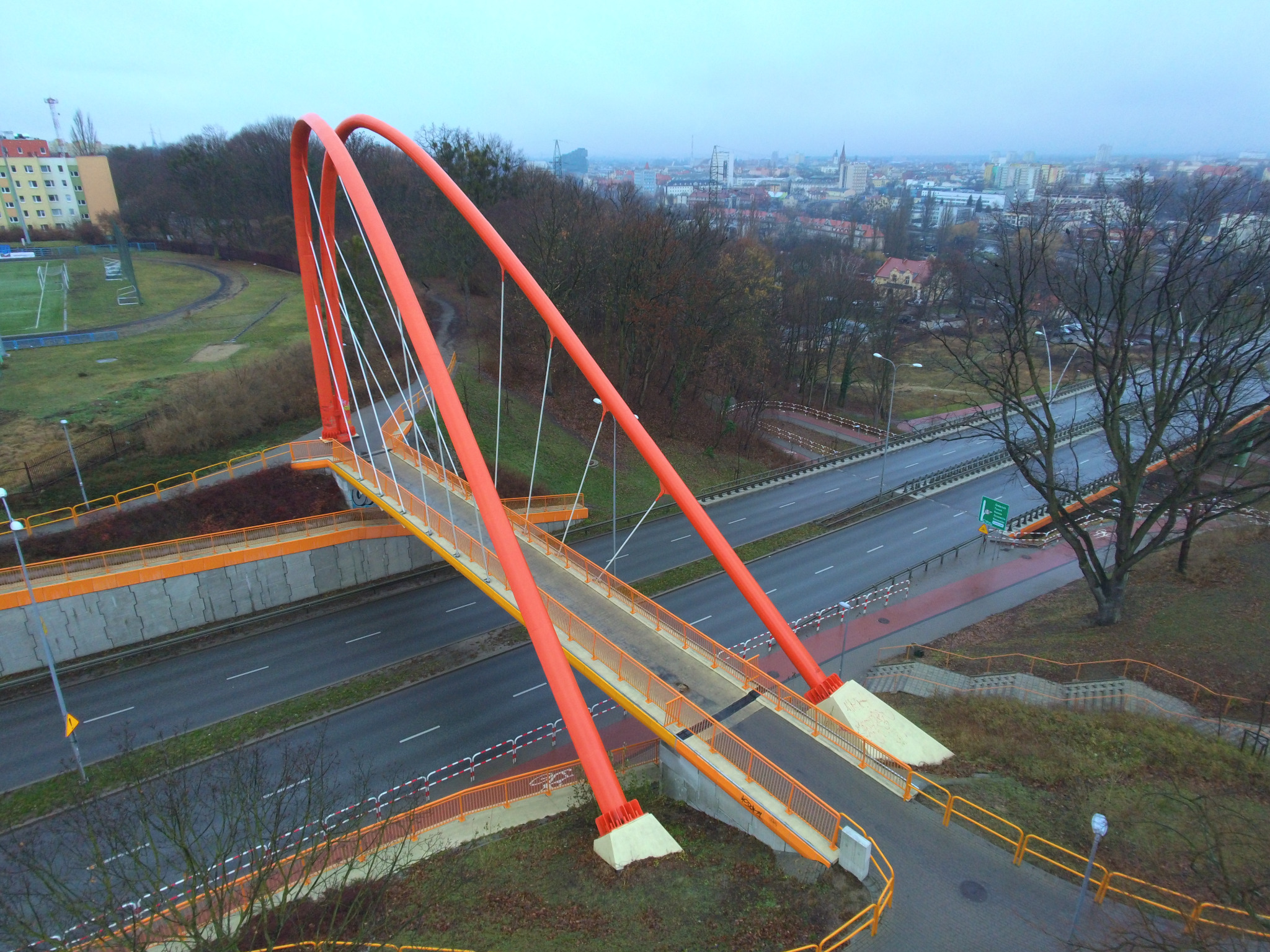 Pomarańczowy most dla pieszych nad ruchliwą drogą, widok z góry w pochmurny dzień, w tle zabudowa miejska.
