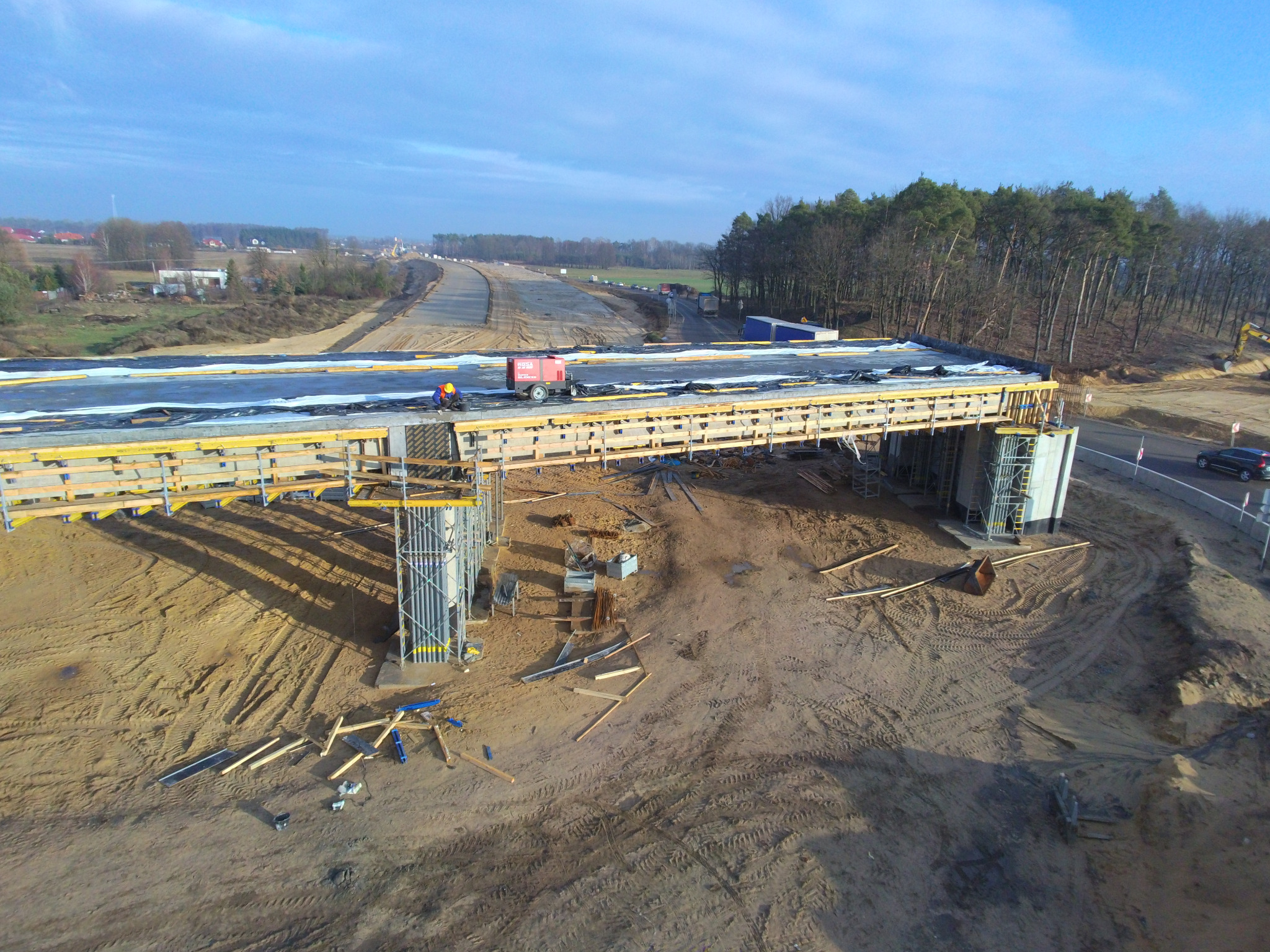 Budowa mostu: widok z góry na konstrukcję z betonowymi filarami i drewnianym szalunkiem, w tle droga w budowie i las, pracownik przy czerwonym agregacie.